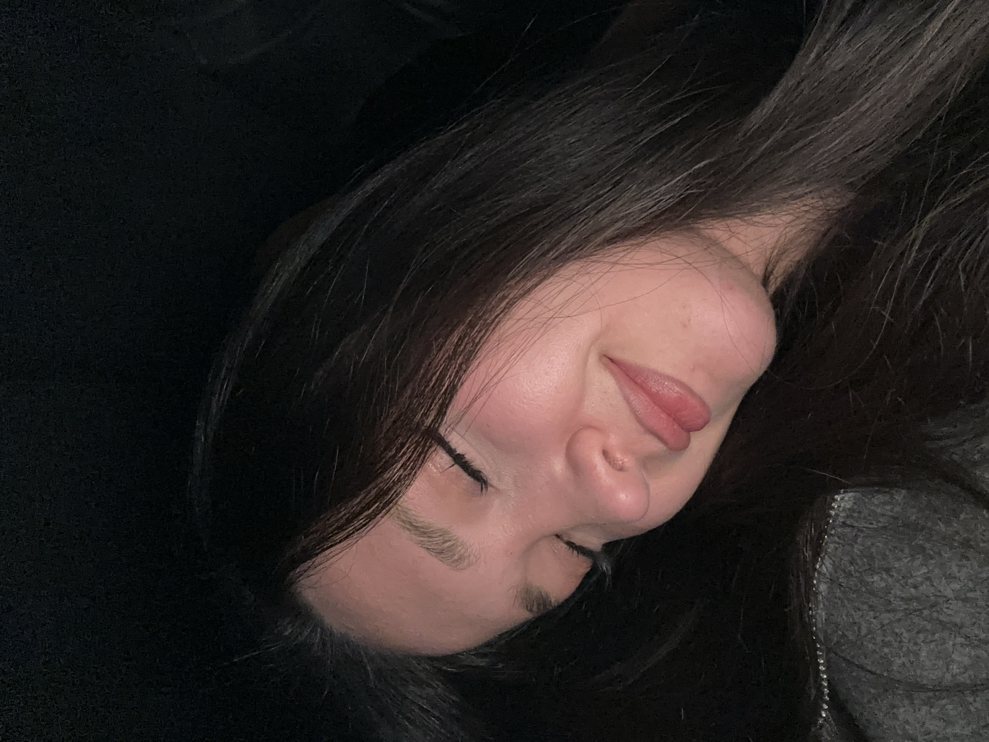 A young woman lies on a dark surface, her head tilted to the side. Her eyes are closed and she has long, dark hair. She is wearing a gray shirt. The background is dark and indistinct. The location is unknown and cannot be determined from the image.