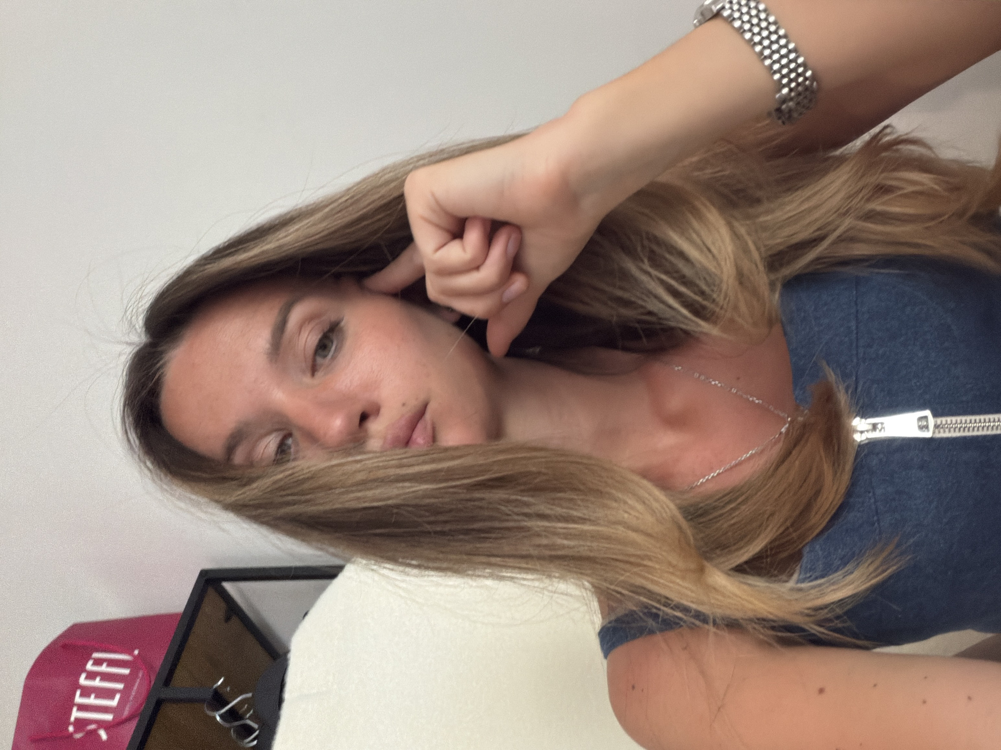 A woman with long blonde hair is shown from a close, angled perspective. She is wearing a blue top with a zipper. Her right hand is touching her ear. Her left arm is visible with a silver watch. A silver necklace is visible around her neck. A pink shopping bag labeled "STEFFI" is partially visible in the bottom left corner. A black metal frame with hanging sunglasses sits beside the bag. The background appears to be a plain white wall. The city and specific location are unknown.