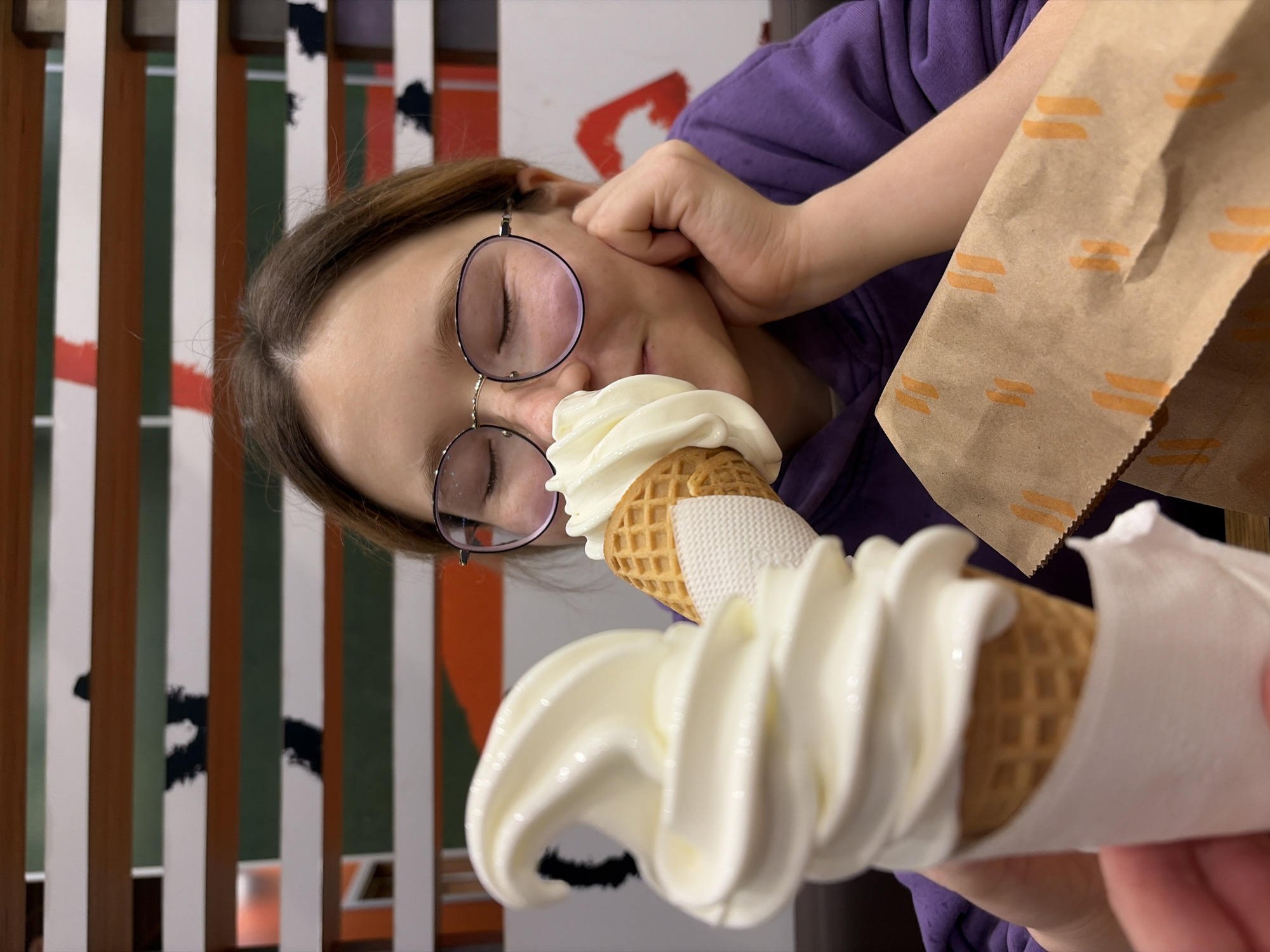 An unidentifiable young person with brown hair, wearing round glasses and a purple hoodie, is pictured in an indoor setting with a blurry background. The person has their eyes closed and appears to be smelling or about to eat a soft-serve ice cream cone held in their right hand. Another ice cream cone is visible in the foreground, also held by an unseen hand. A brown paper bag with orange markings sits on the person's lap. The background features horizontal wooden slats with intermittent green and red abstract shapes.