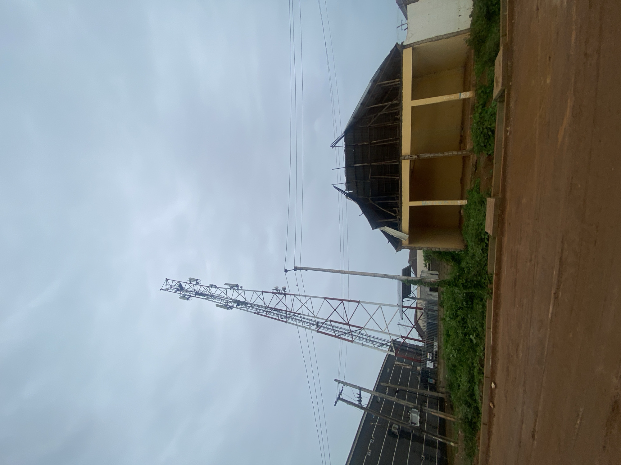 The image shows a street scene in Keffi, Nigeria. In the background, a tall cell tower stands against a cloudy sky. To the right of the tower is a partially constructed or dilapidated building with an open structure and exposed wooden beams. A dark-colored building with horizontal siding is visible to the left of the cell tower. Power lines stretch across the sky, supported by utility poles. The foreground consists of a dirt road with a curb and some overgrown green vegetation. The overall impression is one of an urban environment that may be undergoing development or experiencing some level of disrepair.