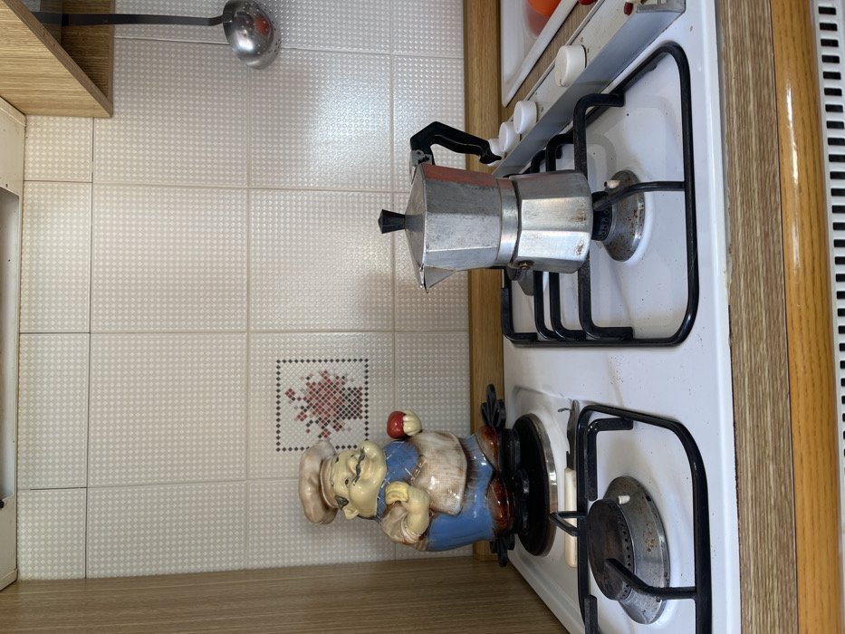 The image displays a kitchen scene, focusing on a countertop and backsplash area, likely in a residential setting in Dosso (Ciappanico), Italy. A white gas stove with four black grates and visible control knobs occupies the right side of the frame. A silver Moka pot, a type of stovetop espresso maker, is placed on one of the front burners, indicating an intention to prepare coffee, although the burner is not actively lit.
To the left of the stove, standing on the wooden countertop, is a ceramic figurine of a jovial chef. The chef wears a white hat, a light-colored vest, and blue pants, and appears to be holding a small red apple in one hand. The backsplash behind the counter is covered with light-colored square tiles featuring a subtle, repeating diamond pattern. Among these tiles, a single decorative tile with a red and black cross-stitch-like floral motif is visible near the bottom center. In the upper left corner, a metal ladle with a red handle or tip hangs, partially obscured by a wooden cabinet or shelf. The stove's burners and grates show signs of regular use. No people are present in the image, and no visible text can be identified. The scene is well-lit, but the specific time of day is not discernable.
