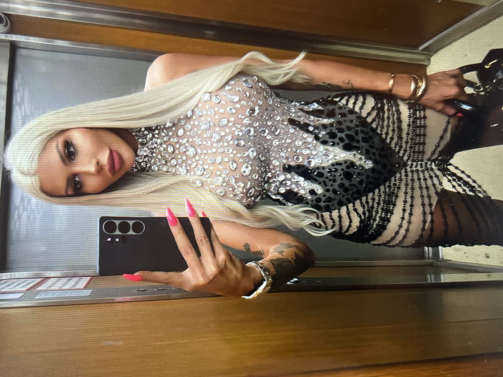 A woman with long blonde hair and bright pink nails stands in an elevator, taking a selfie with her phone. She is wearing a revealing, sparkly dress with black and leopard print accents. She has tattoos on her arms and wears several bracelets on her left wrist. The elevator has wooden-paneled walls and a metallic door. There are no specific details in the image to indicate the location is in the city of Vratsa, Bulgaria.