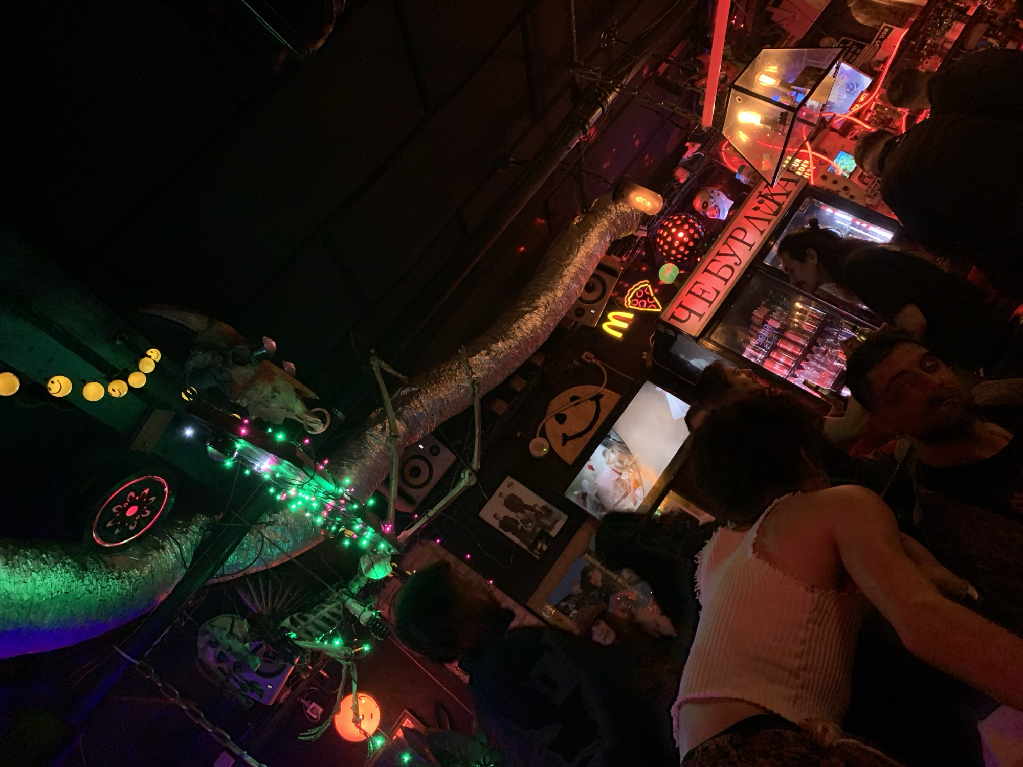 The image depicts a dimly lit interior scene in what appears to be a bar or club in Tel Aviv, Israel. The setting is characterized by eclectic and somewhat haphazard decor. An aluminum duct runs along the ceiling, adorned with string lights and Halloween-themed decorations like skeletons and skulls. Neon signs, including a pizza slice, a McDonald's 'M', and Cyrillic lettering, are visible. Several people are present, including at least one individual with their back to the camera, wearing a white tank top. Other people are near what appears to be a refrigerated display case, possibly interacting with staff. The overall atmosphere is dark, cluttered, and vibrant.