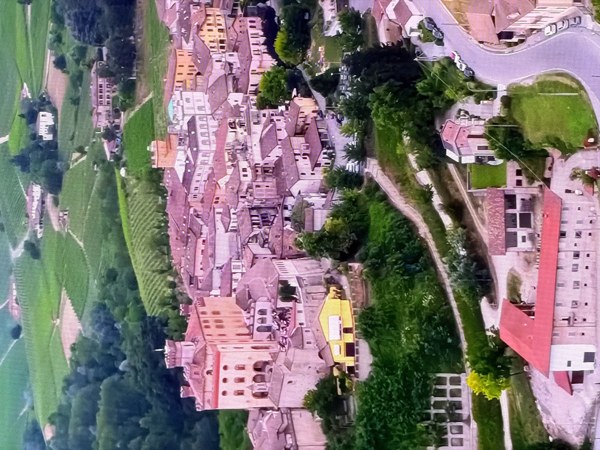 The image displays an aerial view of a village or small town nestled within rolling green hills. The architecture suggests a European setting, with terracotta-tiled roofs and buildings made of stone or stucco. Prominent structures include a large, fortified building resembling a castle or palazzo in a pale pink hue, with a central tower and arched windows. Smaller residential and commercial buildings with varying roof colors and architectural styles are clustered around it. The surrounding landscape is characterized by terraced vineyards and patches of dense green trees. A winding road with several parked cars is visible on the right side of the image, leading into the town. The scene conveys a sense of rural tranquility and historical charm. There is no discernible presence of the city of Chirchik, Uzbekistan.