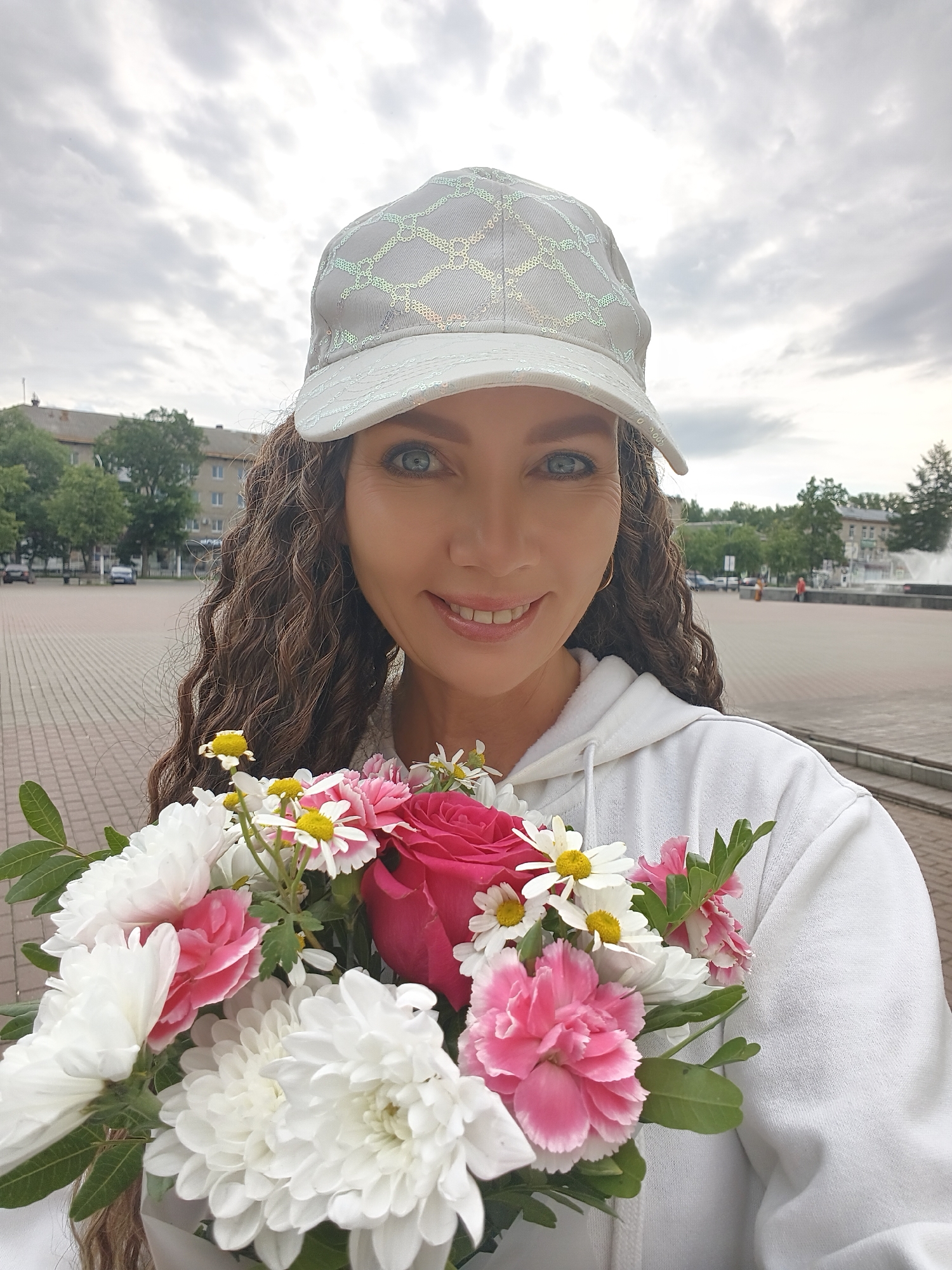 A woman, wearing a white baseball cap with a diamond pattern and a white hoodie, holds a bouquet of flowers. She is standing in an outdoor plaza in the city of Chaikovsky, Russia, as indicated by the buildings, trees, and a fountain visible in the background. The woman's hair is long and curly. The sky is overcast.