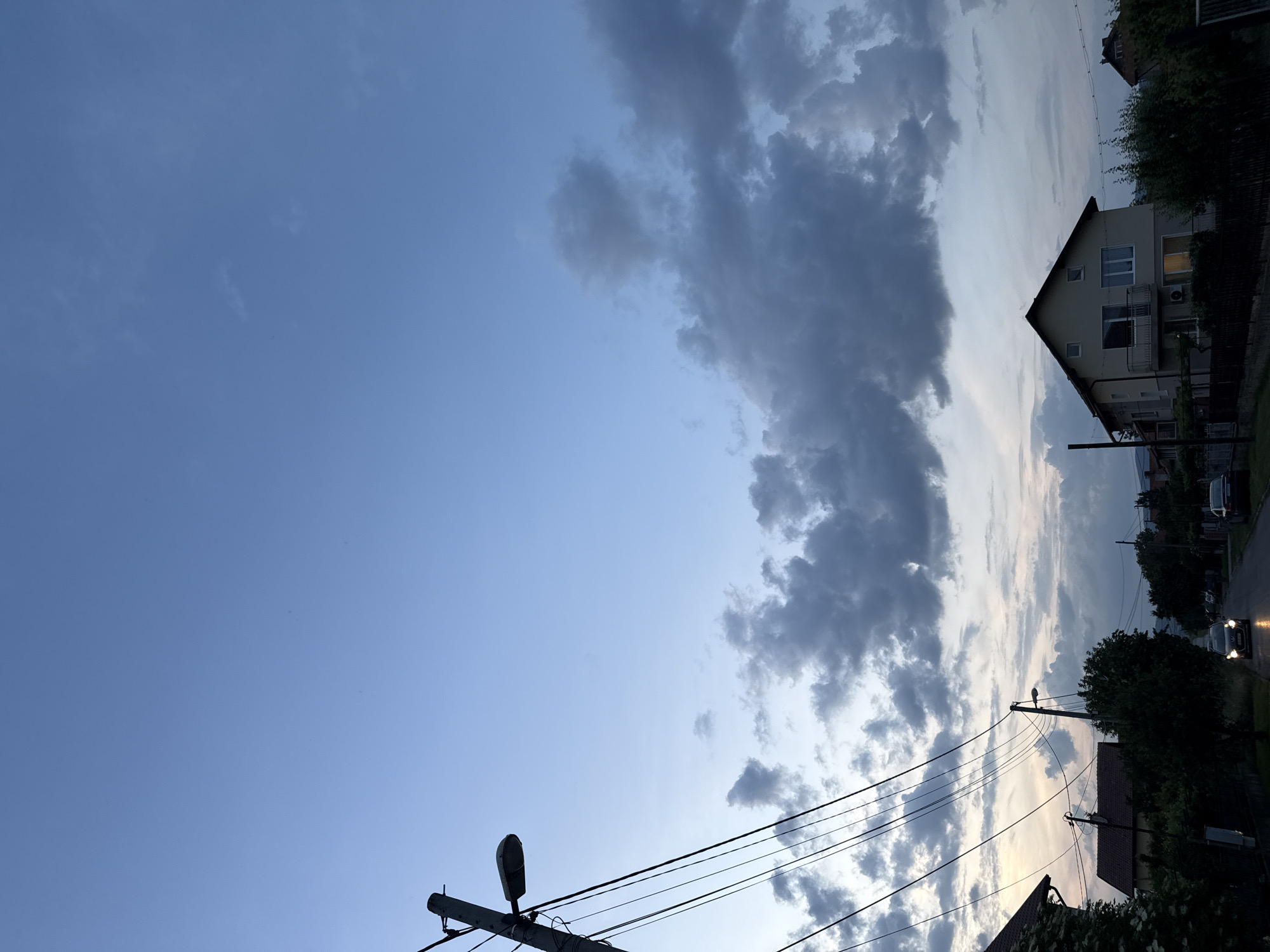 The image depicts a residential street scene in Mirovyane, Bulgaria, captured during late dusk or early night. The sky transitions from a clear, muted blue-grey at the top, to lighter hues near the horizon, where faint remnants of natural light are visible. Overcast conditions are present in the mid-to-lower sky, dominated by irregular, dense dark grey cumulus clouds.
In the left foreground, a concrete utility pole with a dark-colored street light extends upwards, from which numerous black utility wires crisscross downwards across the upper left and mid-left portions of the frame. Partially visible on the far left are the brown-tiled roof of a building and dark green foliage.
A paved road recedes into the distance through the center of the image. A dark-colored vehicle with illuminated headlights is traveling away from the viewer on this road, with reflections of its lights visible on the road surface, suggesting wetness or reflectivity. Another vehicle is parked further down the street on the right. The road is bordered by dense trees and bushes.
On the right side of the image, a two-story residential house with a light-colored facade and a dark-colored pitched roof is prominent. It features multiple windows, some with white frames, and a small balcony on the first floor. A single interior light emits a warm glow from a ground-floor window. A dark fence is visible in front of the house. Other buildings are faintly discernible in the distance, partially obscured by foliage and the low light conditions. No people are visible in the scene. The overall ambiance is quiet, marked by the artificial illumination amidst the fading natural light.