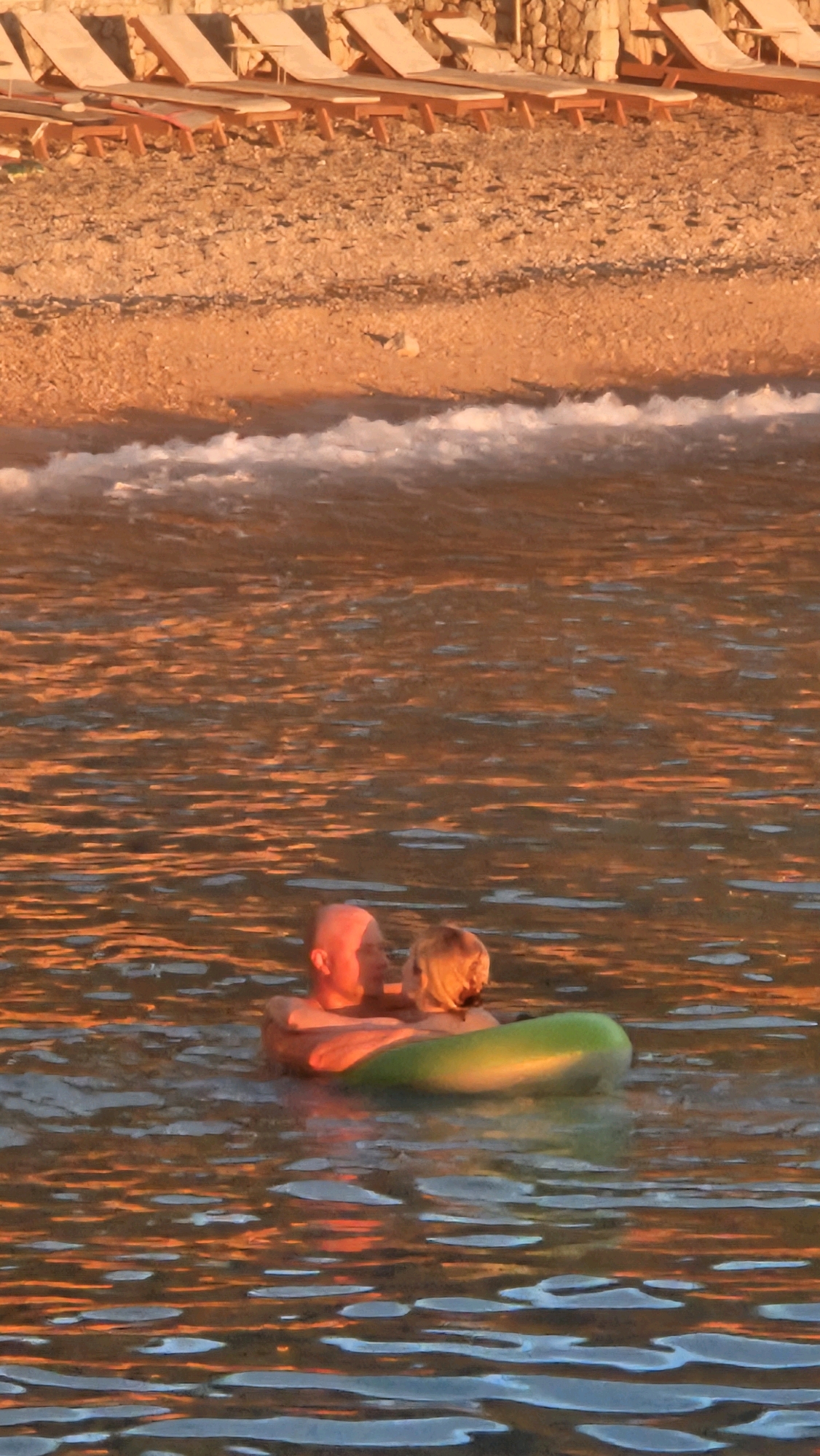 Two individuals, a man and a child, are in the water at sea, floating inside a green inflatable ring. The man, who is bald and has his back to the camera, is looking towards the child. The child, with blonde hair, is facing the man. They appear to be in a calm, shallow sea with sunlight reflecting off the water's surface, creating an orange hue. In the background, there is a beach with several empty lounge chairs set up. The beach is covered in small pebbles and sand, and a stone wall is visible further up the beach. Gentle waves are lapping at the shore. The scene appears to be from a coastal location, possibly a beach resort. The location is identified as Općina Tučepi, Croatia.