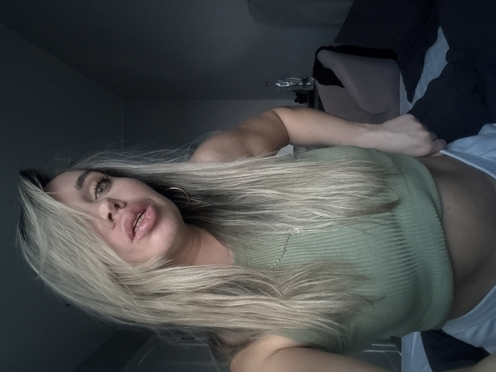 The image displays a female subject in an indoor setting within Votkinsk, Russia. The subject has long, straight blonde hair, parted to the left with strands partially obscuring her left eye. She possesses light-colored eyes and full, slightly parted lips. She wears a light green ribbed crop top, a thin gold chain necklace, and a gold hoop earring in her right ear. Her right hand is positioned at her left hip, grasping the white waistband of what appears to be shorts or underwear, partially revealing her abdomen. The subject is looking directly towards the viewer with a slight puckering of the lips. The environment is dimly lit, with a dark grey wall forming the background. Visible background elements include a partial view of a light-colored, possibly upholstered chair or headboard, a clear plastic bottle with a red cap on a surface, and dark-colored bedding beside white bedding in the lower right and foreground.