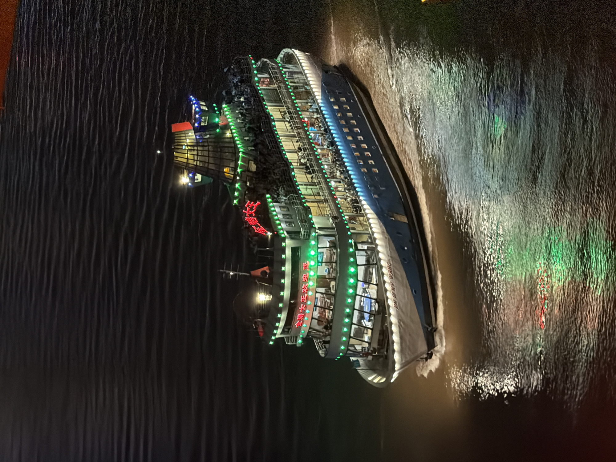 An illuminated cruise ship is navigating a dark body of water, casting reflections. The ship, which appears to be in the Yuzhong District of China, is adorned with numerous green and some red lights along its multiple decks. Many people are visible on the decks, suggesting a crowded passenger vessel. The ship's hull is white and blue, with the name "JIM BENY WANG" visible near the bow. The water is choppy, indicating movement, and the overall scene is nighttime.