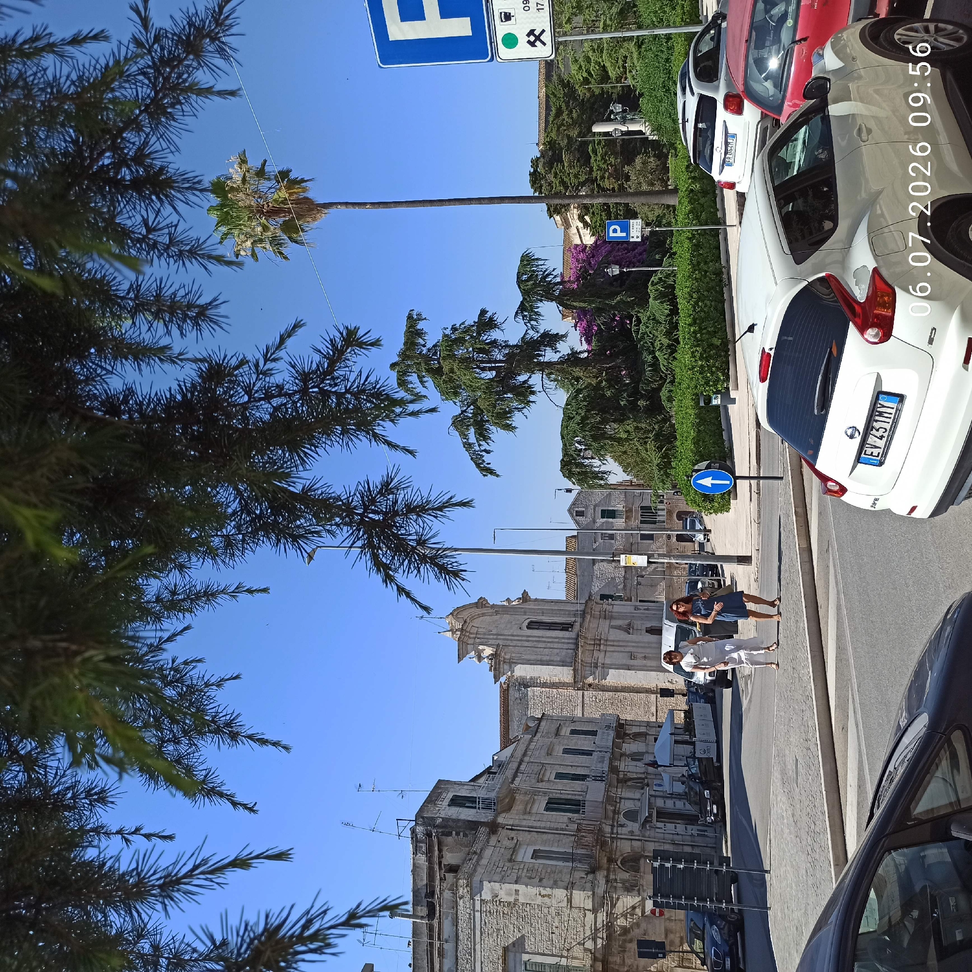In Ruvo di Puglia, Italy, on July 6, 2026, at 09:56, a street scene unfolds under a clear blue sky. The foreground is partially obscured by the dark green needles of pine trees. A white Nissan Juke is parked in the lower right corner, its license plate reading "EV 431MY". To its left, the front of a dark-colored car is visible.
In the mid-ground, a cobblestone street crosses the frame, bordered by a sidewalk. Two women are walking on the sidewalk, one in a white dress and the other in a blue dress. Behind them, a white van is parked. Further back, a dark sedan is parked on the street. Across the street, a row of stone buildings with arched windows and balconies lines the road. A church with a domed roof and a light-colored facade stands prominently in the background.
The environment is characterized by Mediterranean architecture and lush greenery, including palm trees and manicured hedges. Traffic signs are visible, including a blue parking sign with a white "P" and a sign with a white arrow pointing upwards on a blue circular background. The overall impression is of a sunny day in an Italian town.