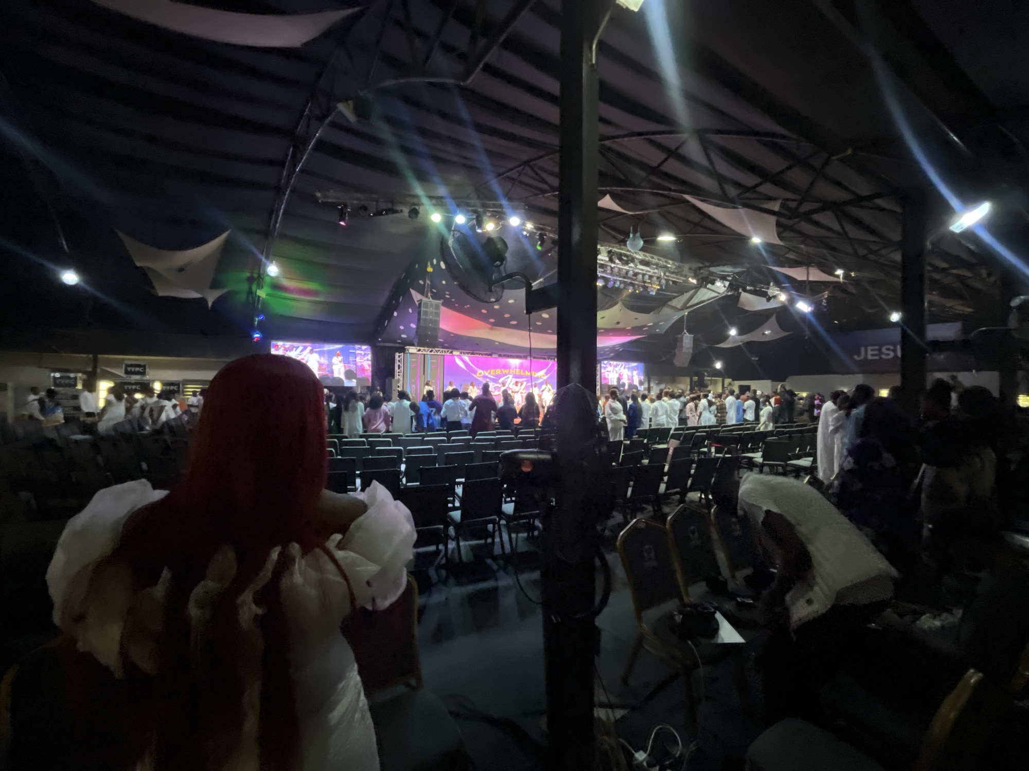 The image shows a large gathering of people inside a building with a high, dark ceiling supported by metal beams. Decorative white fabric is suspended from the ceiling. A stage is visible in the background with screens displaying content and people standing on it. Rows of empty chairs are arranged facing the stage.
In the foreground, there are two people. One is wearing a red head covering and a white garment with decorative white ruffles. The other person kneels on the right side of the frame, their head covered in white fabric.
Based on the “JESI” sign on a blue banner near the stage, it is highly probable that the location is a church service or a religious gathering. Ado, Nigeria is not directly identifiable in the scene.