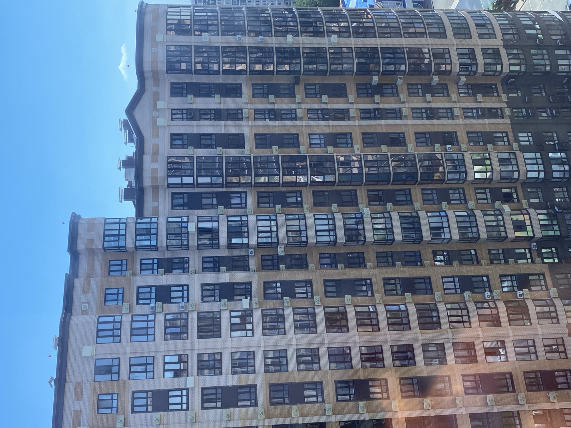 An exterior, eye-level, medium shot captures a tall, modern apartment building on a clear, blue-sky day. The building, constructed with light-brown brick and beige accents, features numerous large, dark-framed windows and balconies. The windows reflect the sky, creating a glazed appearance. Air conditioning units are visible on some of the balconies. The building has a curved section on the right side. The surrounding environment is not visible beyond the building itself.