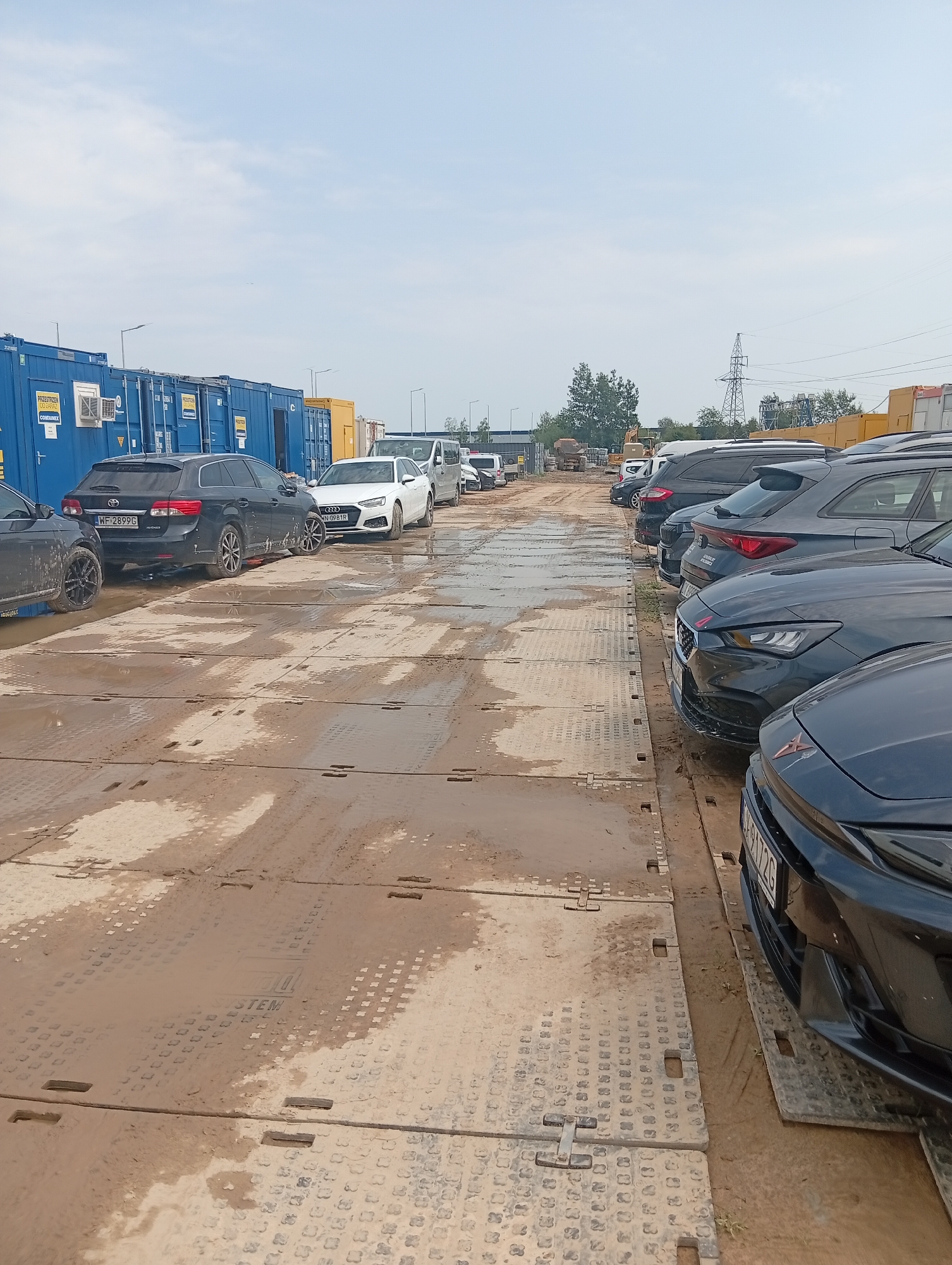 A linear perspective photograph taken outdoors on a cloudy day shows a row of parked cars on a muddy, temporary-looking surface composed of interlocking metal plates, likely in a construction or storage area. The vehicles, primarily SUVs and sedans, are parked in a line, facing towards the right side of the frame. Some of the cars are dark-colored, while others are white. In the background, several large, blue shipping containers are visible, suggesting a logistics or industrial setting. A yellow portable structure and some construction equipment, including a crane, are also present in the distance. The ground surface is wet and muddy, with puddles and dirt accumulated on the metal plates. The sky is overcast, with a few wisps of clouds. The overall scene depicts a practical, utilitarian outdoor space.