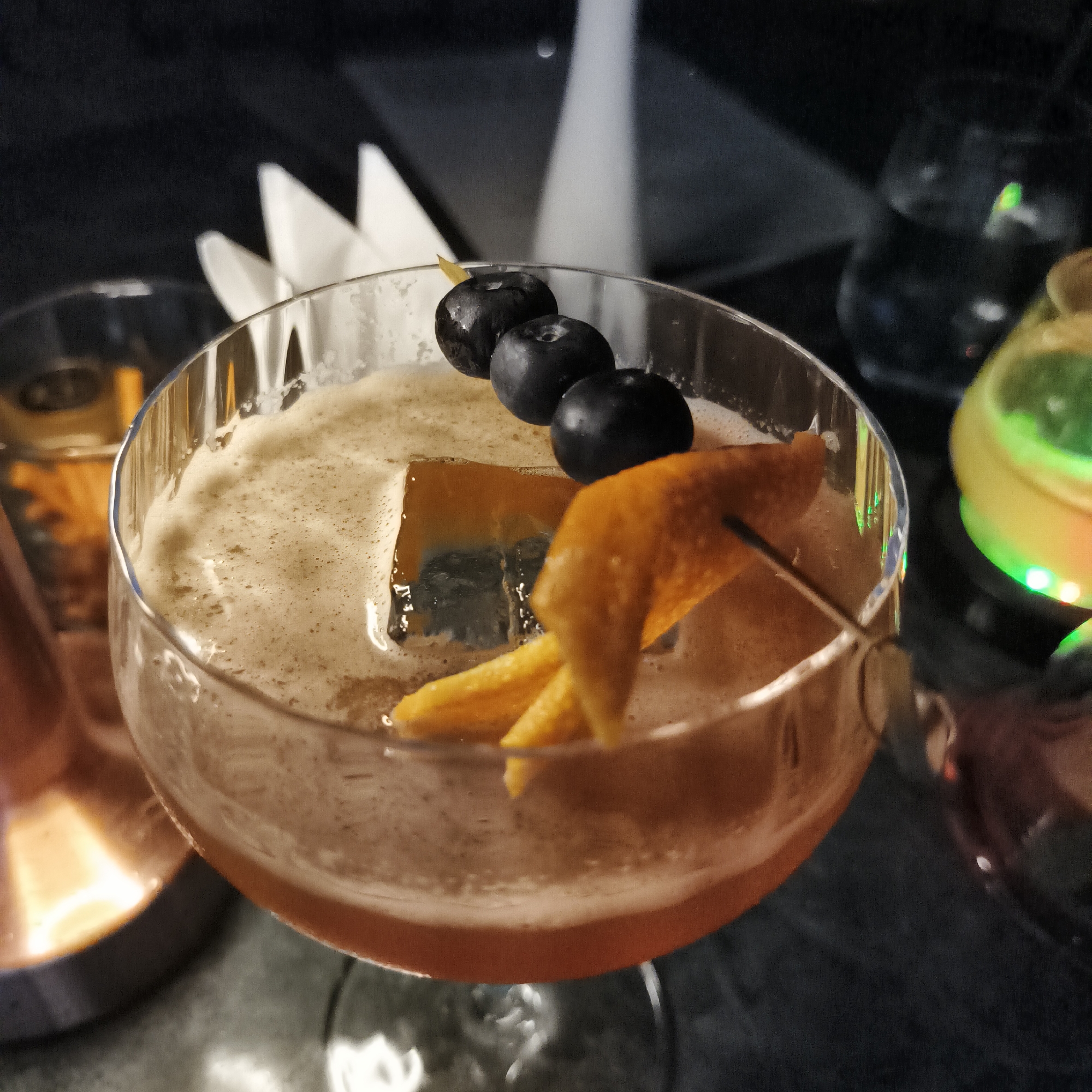 This media file captures a close-up shot of a cocktail in a coupe glass, set in a dimly lit bar or restaurant environment. The cocktail features a frothy head, a reddish-brown liquid, and a large, square ice cube. It is garnished with a twisted orange peel, neatly placed on the rim, and three dark blueberries skewered together, resting on top of the drink.
In the background, out of focus, are several elements that suggest a sophisticated setting. To the left, part of another glass with an orange garnish is visible. A white, folded paper object, possibly a napkin or menu, stands upright behind the main cocktail glass. On the right, a glass containing a glowing green light can be seen, adding to the ambient lighting. The overall background is dark, with subtle reflections on what appears to be a dark tabletop. The low light conditions indicate that the scene is set in the evening or at night. The image conveys a relaxed and elegant atmosphere, typical of a cocktail bar in a city like Bangkok, Thailand. There is no visible text in the image.