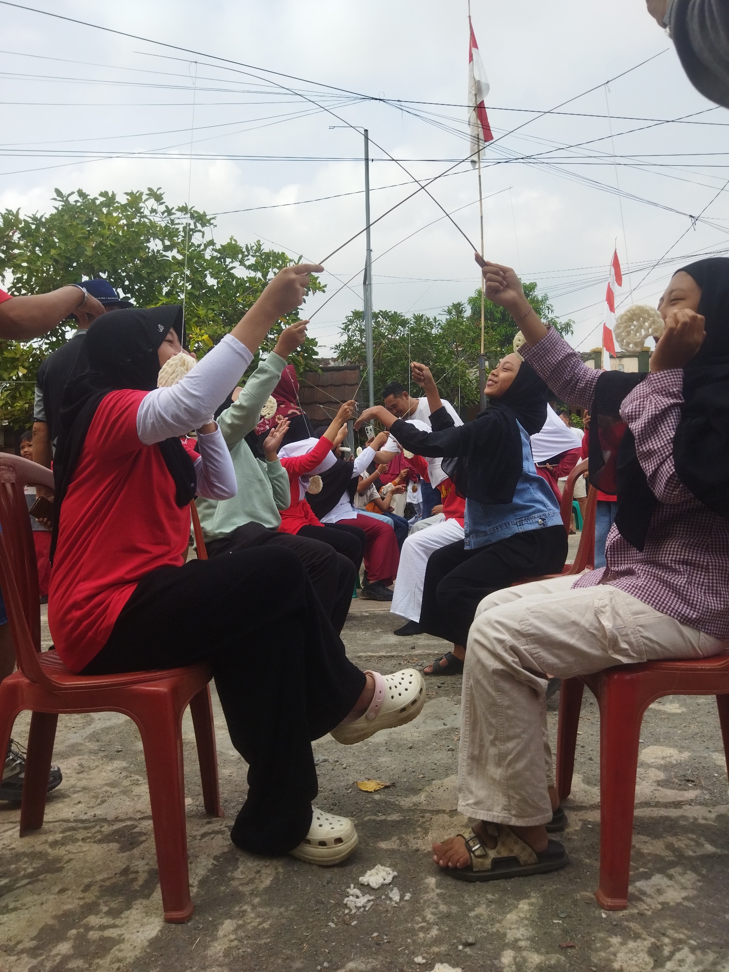 In Banjar, Indonesia, a group of people are participating in a cracker-eating competition. The competition involves individuals sitting on red plastic chairs, holding a cracker attached to a string with their mouths. The cracker is suspended above them, and they attempt to eat it without using their hands. The event appears to be outdoors, with trees and a cloudy sky visible in the background. Indonesian flags are also visible, suggesting a national celebration or holiday. The participants are dressed casually, and some are wearing hijabs. The ground is paved with concrete.