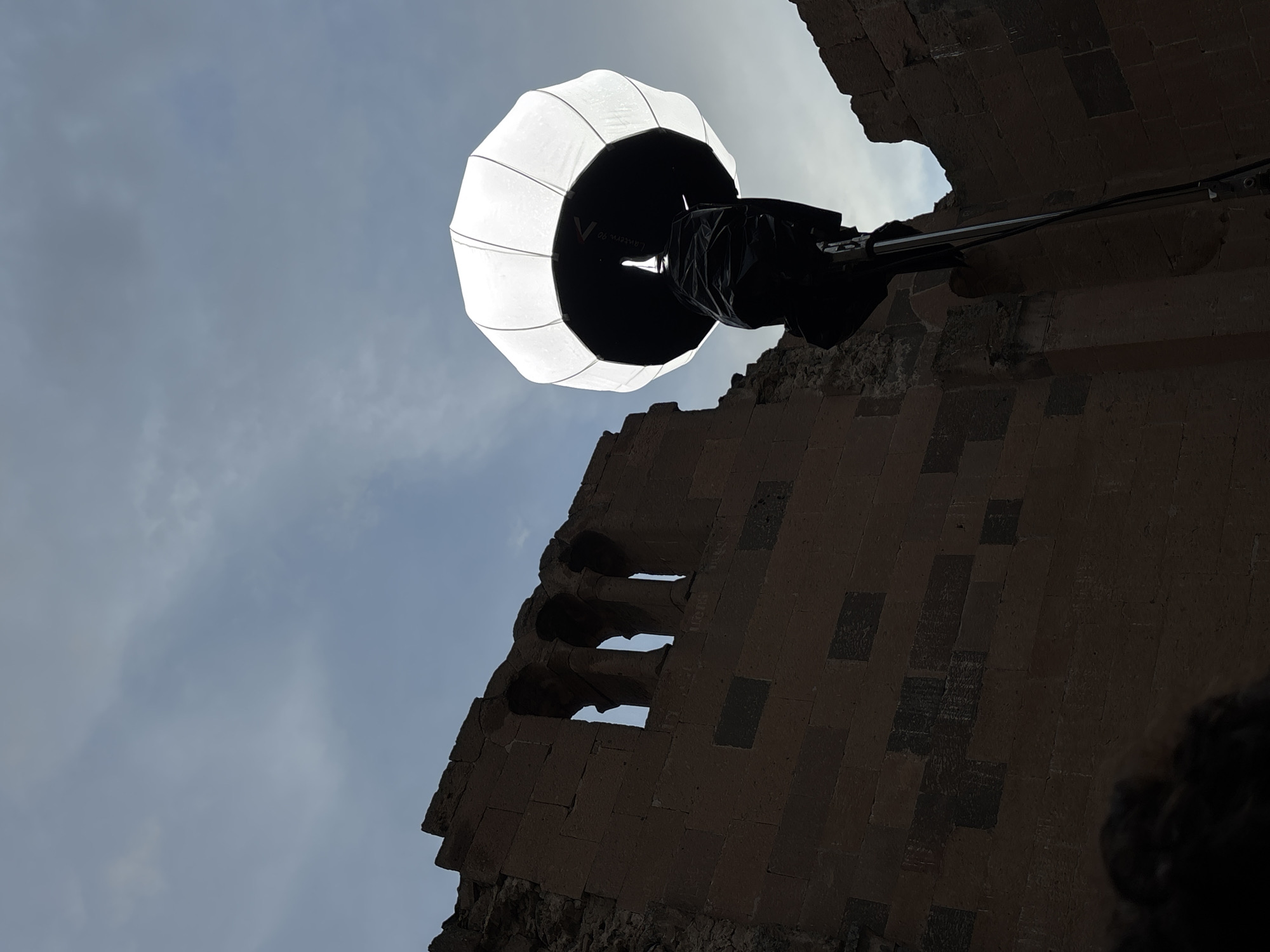The image captures a low-angle view of a building in Anipemza, Armenia, taken in daylight. Dominating the upper part of the image is a bright, lantern-like light diffuser held in place by a dark arm extending from the building. The building itself is constructed from stone blocks, with a series of three openings or windows visible in the structure. The sky is visible through the openings and around the structures, revealing a cloudy day. A small portion of a person or object is visible in the bottom right corner.