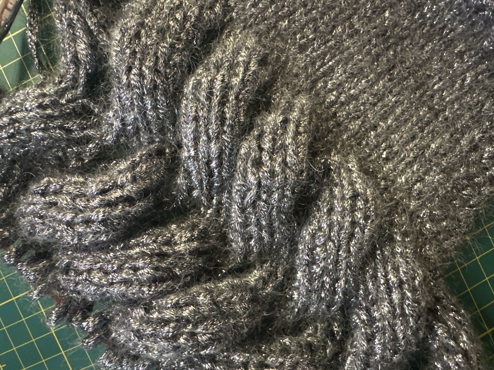 A close-up, overhead shot shows a textured, dark gray knitted fabric with silver metallic threads woven throughout, creating a shimmering effect. The fabric is draped and folded, revealing a cable knit pattern in some areas and a looser, textured knit in others. The background is a green cutting mat with yellow grid lines. No people or specific city information are visible in the image.