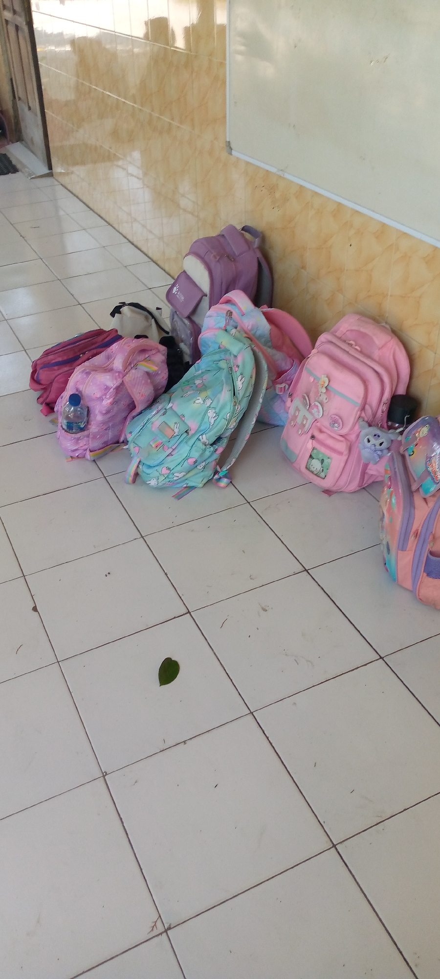 The image depicts a hallway with tiled floors and a wall covered in light brown tiles. Several backpacks are scattered on the floor, leaning against the wall. One backpack is purple and gray, another is pink with unicorn and rainbow patterns, and a third is a lighter pink with a unicorn design. To the right, a larger pink backpack with a stuffed animal attached is visible, along with part of another backpack. A green leaf rests on the floor in the foreground. The scene appears to be in a school or educational institution in Pacing, Indonesia, given the context of backpacks.