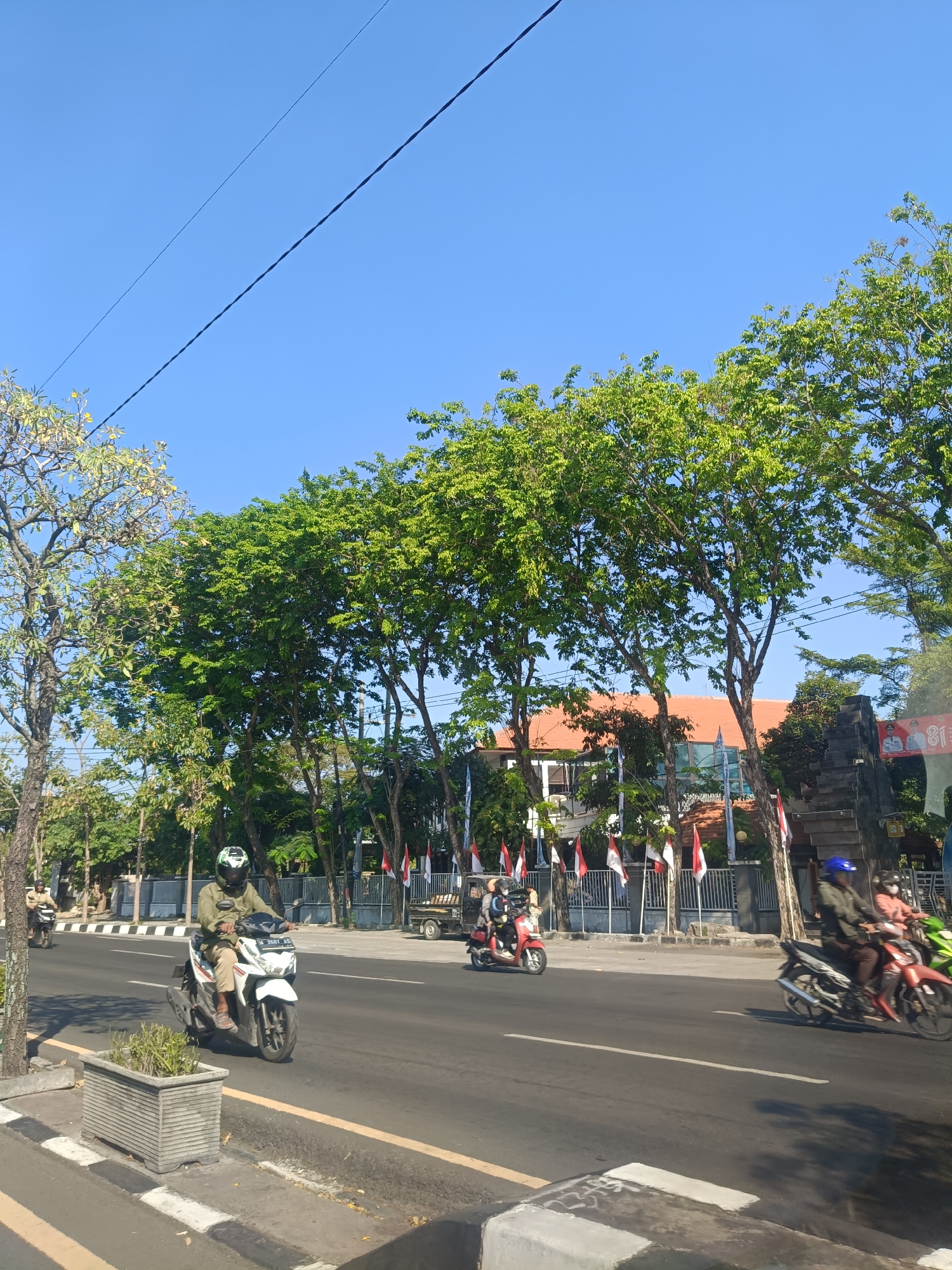 This is an eye-level, medium shot of a street scene in Dahanrejo, Indonesia, under a clear blue sky. In the foreground, a white scooter with a rider wearing a green jacket and helmet is traveling on the right side of the road. The scooter has a license plate that reads "N 2581 LG". To the left of the scooter is a grey concrete planter box with a small green plant. The road has a yellow line separating the lanes.
In the background, several trees with lush green foliage line the street. Behind the trees, a building with a red-tiled roof is visible, along with a stone structure adorned with flags. Various other vehicles, including motorcycles and a small truck, are seen on the road. The overall scene depicts a bright, sunny day in a residential or commercial area of Dahanrejo.