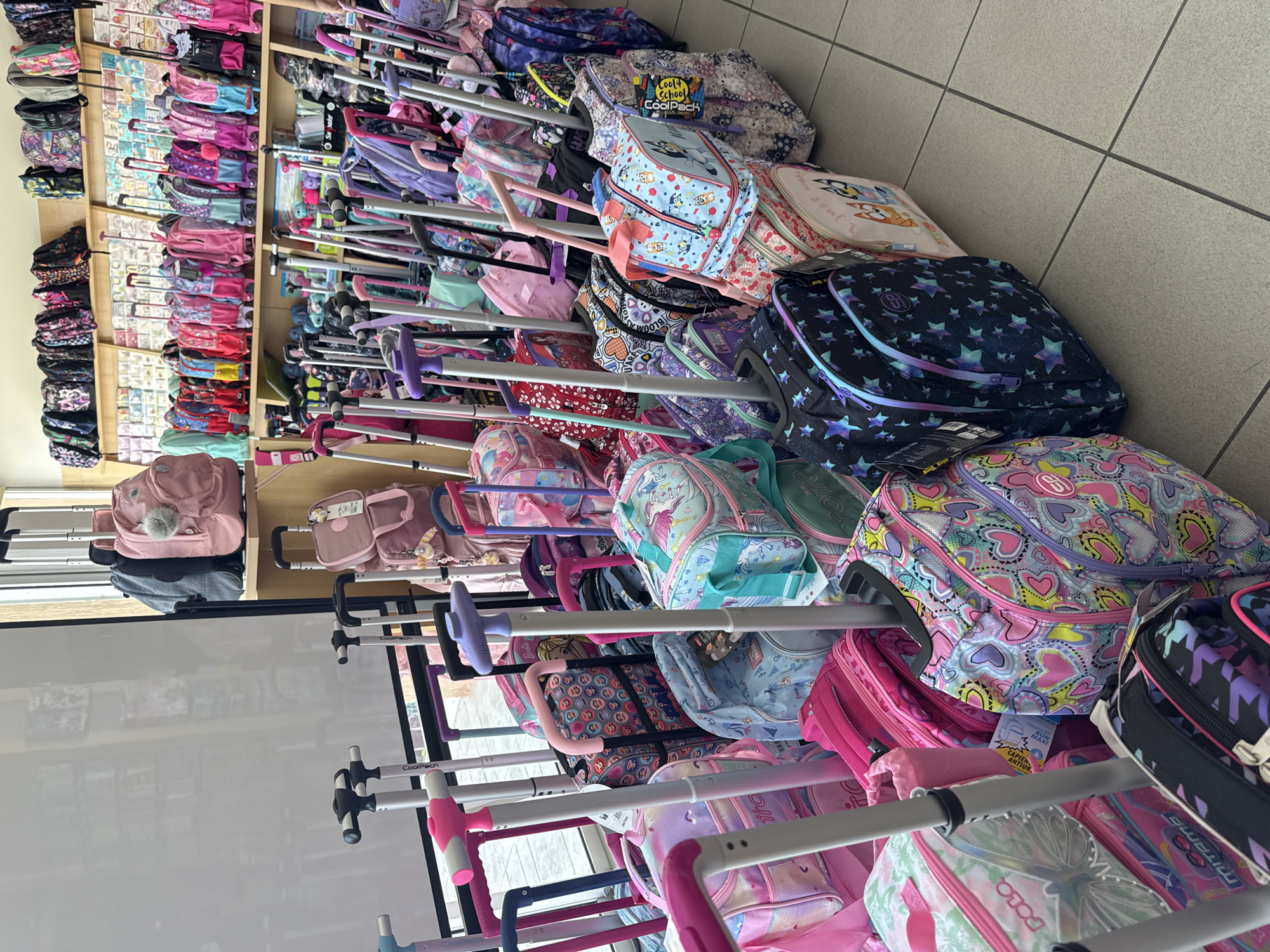 In Agios Athanasios Municipality, Cyprus, a retail store displays a large collection of wheeled backpacks and luggage. The scene is dominated by colorful children's backpacks, many featuring popular cartoon characters like Bluey, and various patterns such as hearts, stars, and unicorns. The items are neatly arranged on shelves and in aisles, with many wheeled backpacks standing upright, ready for purchase. The store's lighting is bright, illuminating the vibrant designs of the merchandise. The floor is tiled in a neutral gray color, and the background reveals shelves stocked with more items, suggesting a well-organized retail environment. There are no people visible in the image, but the presence of such a large assortment of children's products indicates a store catering to families and students.