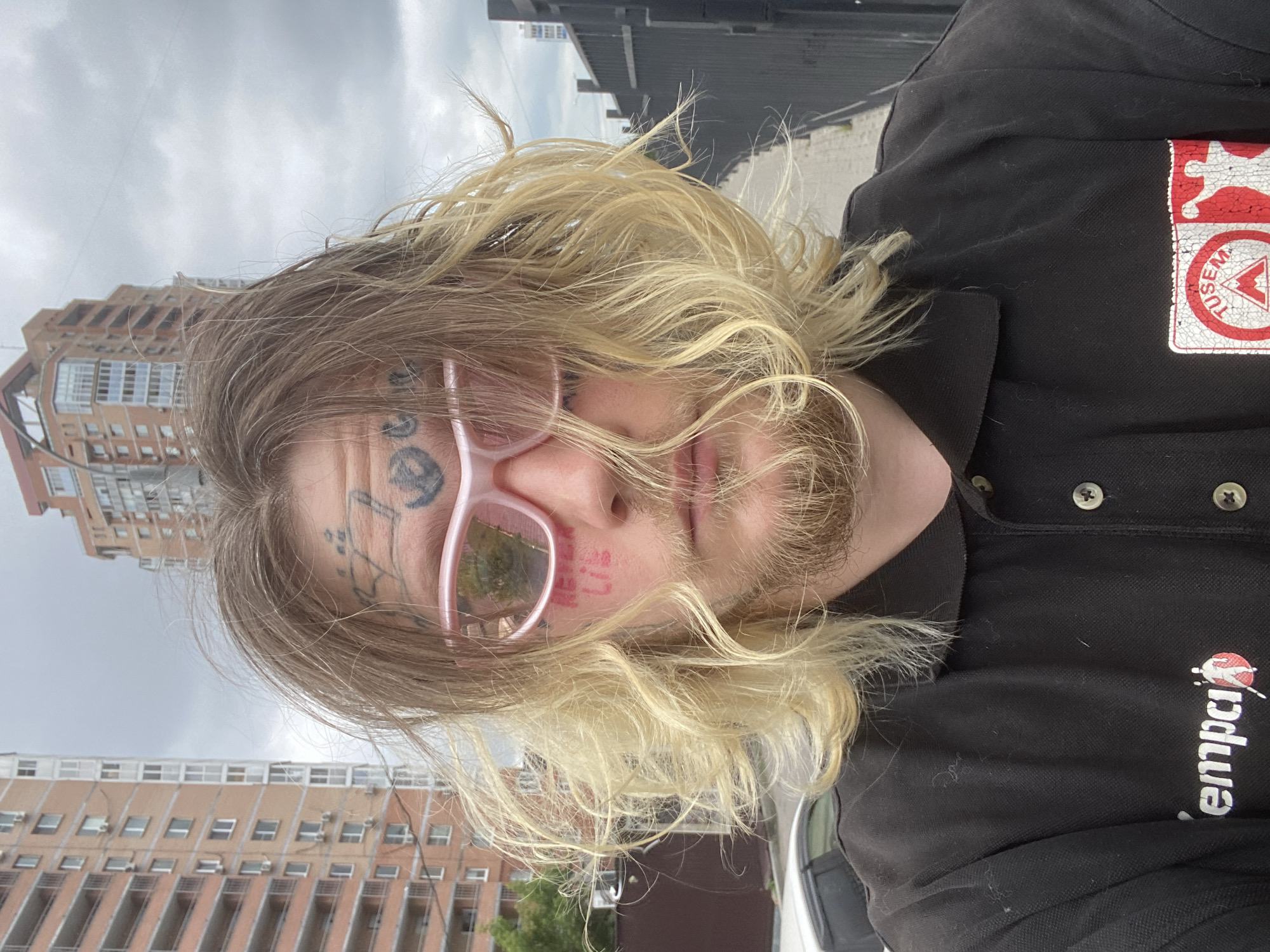 A close-up, front-facing selfie of a person with blonde, unkempt hair, a beard, and facial tattoos. They are wearing pink-tinted sunglasses and a black polo shirt. Red lettering on their cheek appears to read "NEVER LIE." The background shows a cloudy sky, a tall brick apartment building, and a black metal fence. The image appears to have been taken outdoors, possibly in an urban setting. Without more information, it is impossible to determine the exact location within Khabarovsk, Russia.