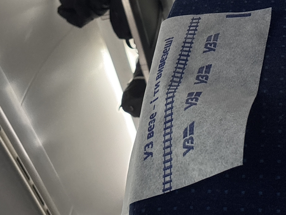 A headrest cover in a train car seat is seen. Printed on the cover in blue ink, are the words "УЗ ВЕЗЕ - I ТИ ВИВЕЗЕШ!" which translates roughly to "UZ takes - and you take out!" along with a line of railway tracks and the UZ logo multiple times. The cover is slightly crumpled and affixed to a dark blue patterned seat. The train car walls and overhead storage compartments are visible in the background.