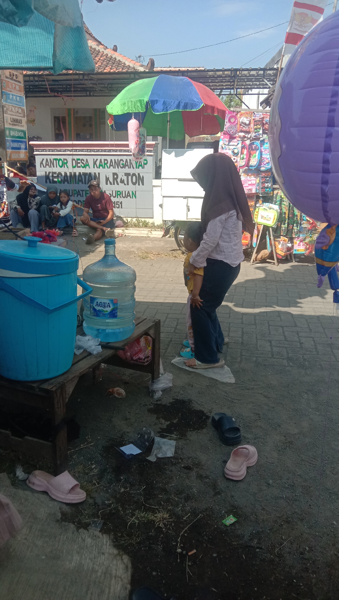 In front of a building with the text "KANTOR DESA KARANG ANYAR KECAMATAN KRATON KABUPATEN SURABAYA" visible, a woman wearing a dark hijab and a checkered shirt is holding a young child. The child is wearing a yellow shirt and patterned pants. They are standing on a piece of white plastic. To their left, a large blue cooler and a large bottle of Aqua water are placed on a wooden stand. Behind them, several people are seated, and a colorful umbrella is open. To the right, a stall displays various items, including bags and toys, and a large purple balloon is visible. The ground is paved with interlocking bricks and appears wet in some areas, with scattered trash. Several pairs of flip-flops are on the ground. The scene appears to be outdoors on a sunny day.