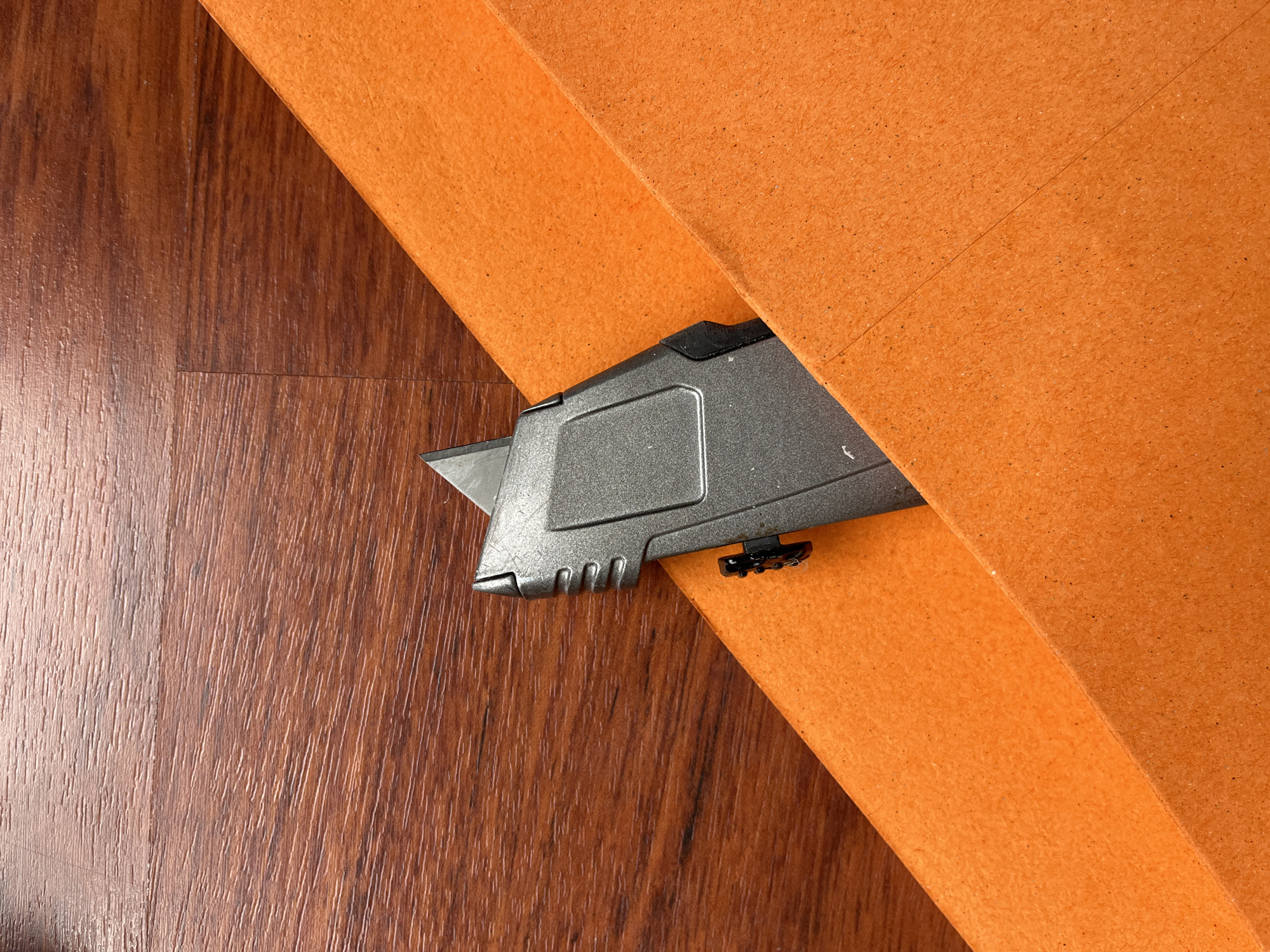 The image shows a utility knife, partially visible, cutting into a piece of orange material on top of what appears to be a wood-patterned floor. The scene consists of these three elements: the utility knife, the orange material, and the flooring. There are no people visible in the image. The image does not provide information to determine if it is in the city of Aurora, United States of America.