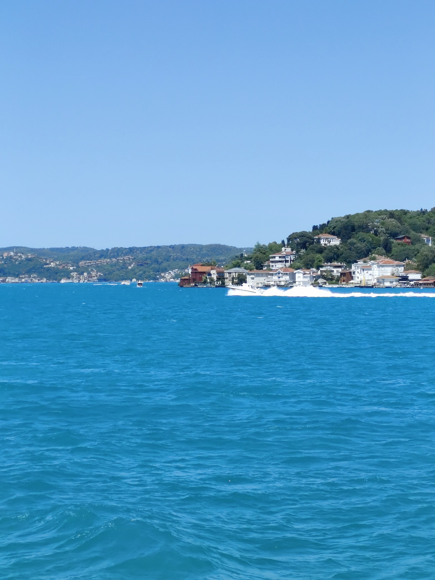 The image depicts a view of the Bosphorus Strait in Sarıyer, Turkey. The foreground is dominated by the turquoise waters of the strait, with gentle ripples visible on the surface. Along the horizon, a densely forested hillside is dotted with white buildings and villas, indicating residential development. Some boats are on the water and a speed boat is creating a white wake along the shoreline. The sky above is a clear, cloudless blue.