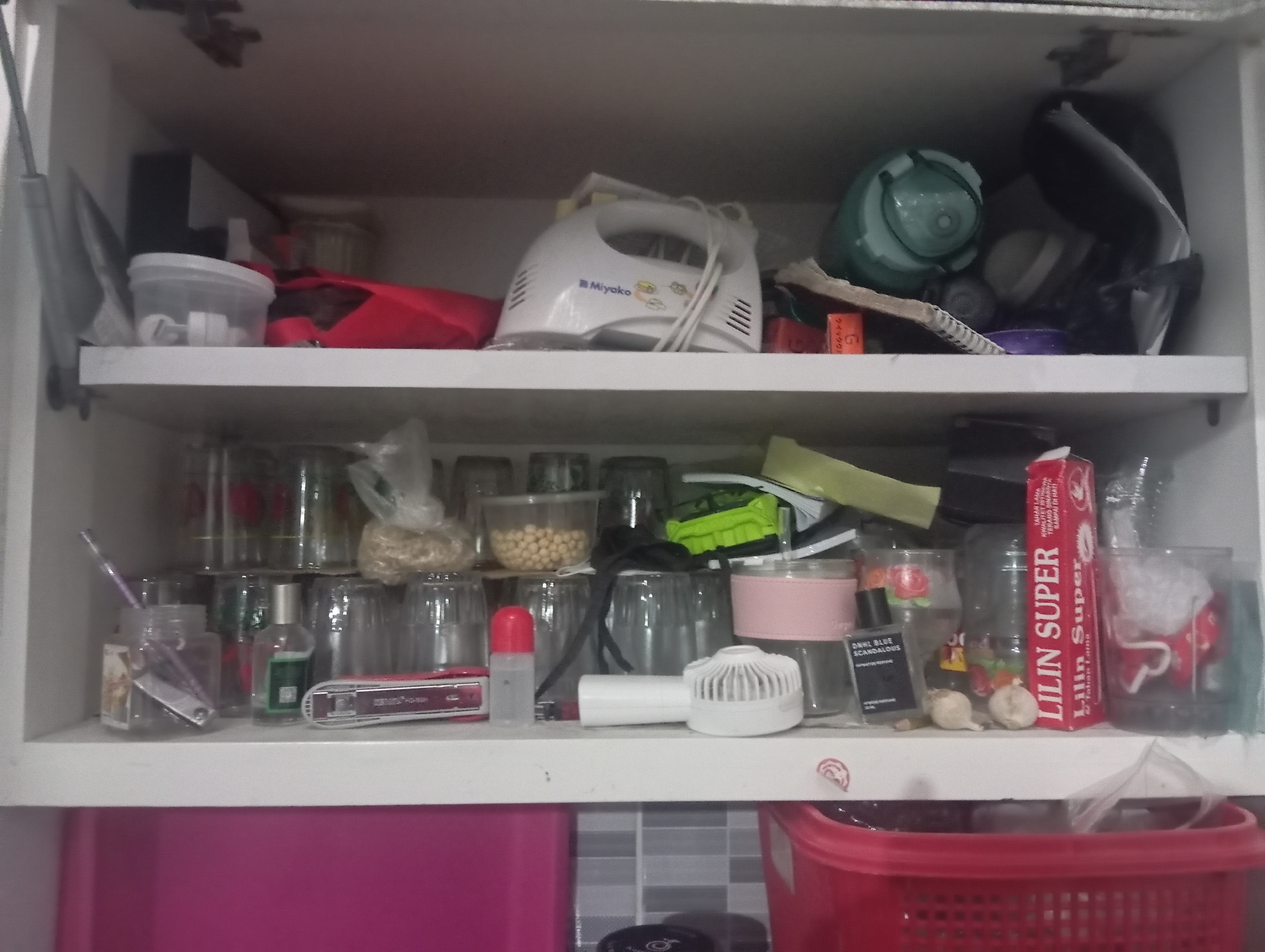 The image depicts an open, white, two-tiered shelf unit, possibly a kitchen cabinet, located in Kutasari, Indonesia. The top shelf contains a white Miyako brand electric mixer, a red bag, and several green and black objects. A green plastic container with a lid is also visible. The bottom shelf is filled with various items including numerous glass containers, some with contents like small yellow spheres. There is also a pink stapler, a small white fan, a bottle of what appears to be perfume or cologne, and a red box with "LILIN SUPER" printed on it. A small, clear plastic bottle with a red cap is also present. The overall environment appears to be a domestic storage space, likely in a kitchen or pantry. The lighting is somewhat dim, suggesting an indoor setting. No people are visible in the image.