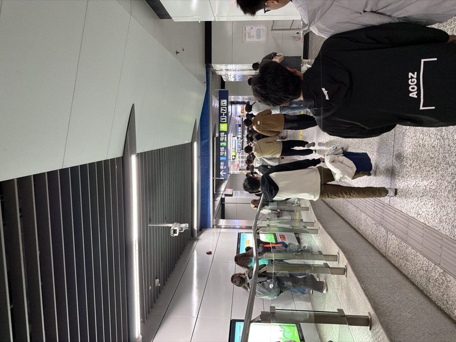An indoor scene depicts a brightly lit, modern subway station concourse or passageway in Yuhuatai District, China. The environment features a dark gray slatted ceiling with linear white fluorescent or LED light fixtures, and white flat panel walls. A white security camera is mounted overhead on the left. The floor is composed of light gray terrazzo-style tiles, with tactile paving strips visible in the foreground.
Approximately 20-30 individuals, predominantly young adults, are present. Most are observed walking away from the camera towards a junction in the midground, suggesting movement towards platforms or exits. In the immediate foreground, the back of a person wearing a black hoodie with "AOGZ" printed in white on the back is visible on the right. To their left, another person, also seen from behind, wears a white and black hoodie, brown trousers, and white sneakers, carrying a blue bag containing white fabric in their left hand.
On the left side of the scene, a raised platform is separated from the main walkway by a glass barrier with stainless steel posts and a top rail. Behind this barrier, several individuals are standing or walking. Digital screens displaying green graphics are embedded in the wall behind them, partially obscured. A red fire alarm pull station is mounted on the left wall. In the midground, a prominent blue overhead sign with white text and arrows directs passengers to "Line 1" and "Line 3," and indicates exits "1-7" in both Chinese and English. People in the background are densely clustered, navigating the passageway. No direct interactions between individuals are discernible; all subjects appear to be in transit.