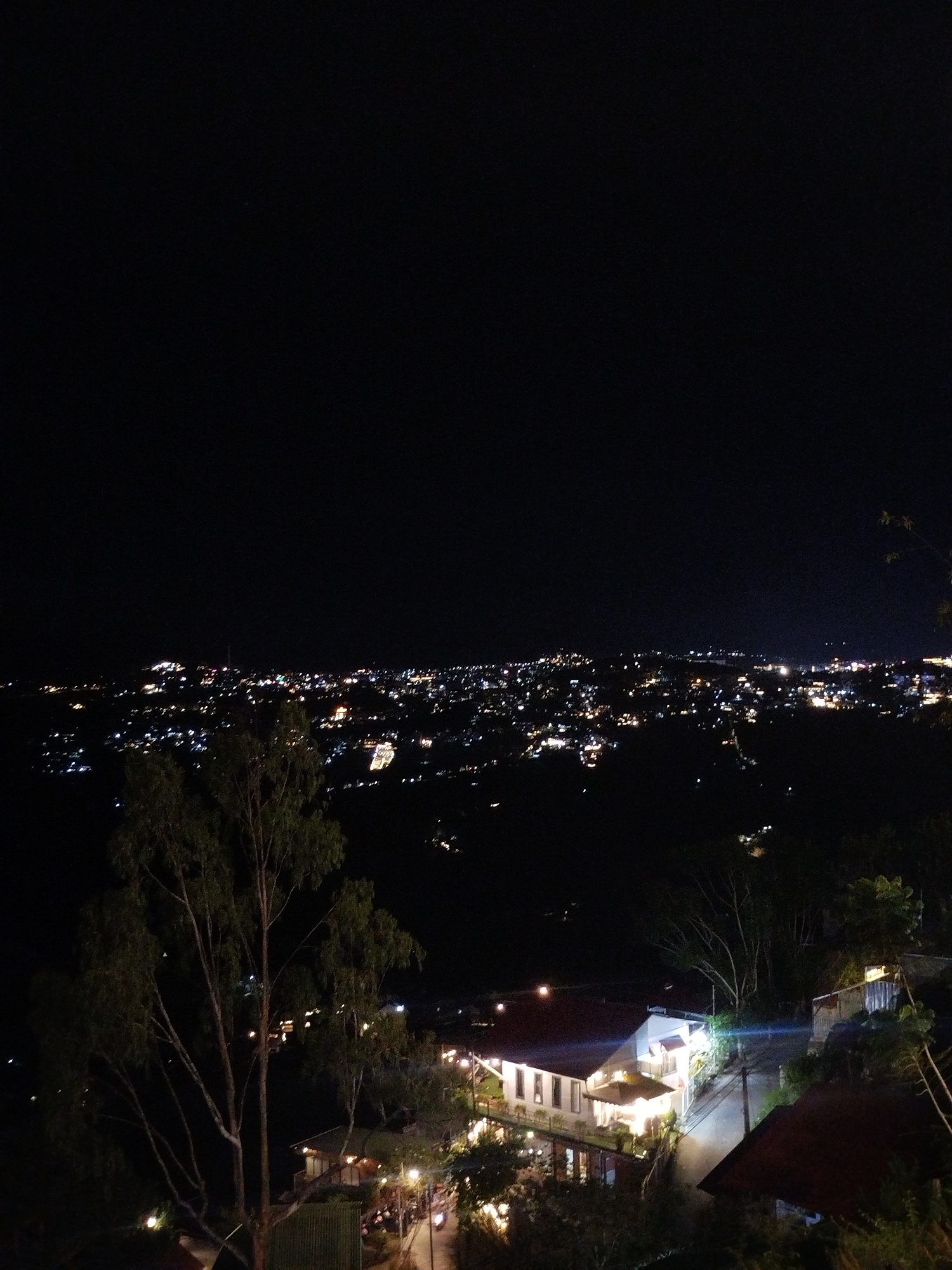 The image depicts a nighttime scene in Xuan Truong, Da Lat, Vietnam. The cityscape is visible in the distance, illuminated by numerous lights. In the foreground, there's a building with well-lit exteriors and surrounding landscape featuring trees. The sky is dark and unobscured.