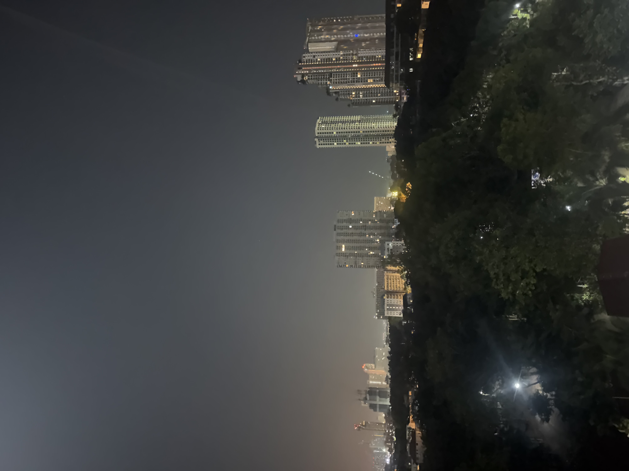 This night photograph presents a panoramic view of Pattaya City, Thailand, captured from a high vantage point. The left side of the frame features a vast expanse of dark, hazy sky, indicative of either atmospheric conditions or distant light pollution.
The central and mid-ground is dominated by a bustling urban landscape, characterized by numerous modern high-rise buildings. Many of these structures, likely residential or commercial towers, display illuminated windows, casting a warm glow across the scene. The buildings are varied in height and design, stretching into the distance where the city lights merge into a diffused, shimmering horizon line.
To the right, a dense concentration of dark green trees and foliage fills the foreground and mid-ground, providing a stark natural contrast to the concrete structures. These trees are largely silhouetted against the ambient city lights, though some interior light sources, possibly streetlights or lights from buildings behind them, are visible peeking through the canopy.
No explicit human activity or discernible figures are present, suggesting a quiet, static moment in the city. The time of day is clearly night, evidenced by the dark sky and extensive artificial illumination. The hazy atmosphere diffuses the city lights, creating a soft, almost ethereal glow. There is no visible text in the image. The scene captures the sprawling, illuminated beauty of a tropical city at night.