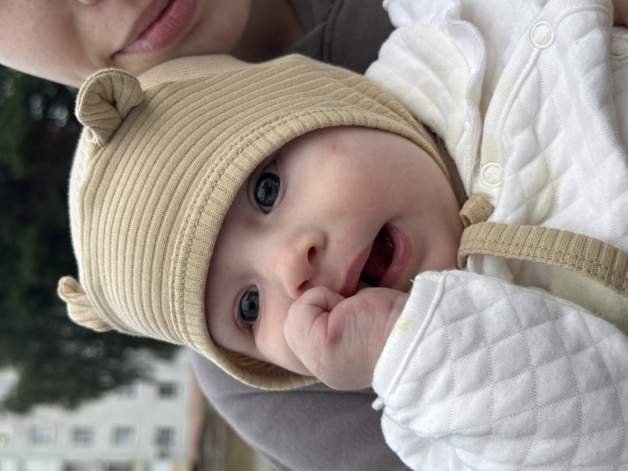A close-up shot captures a baby wearing a tan, ribbed bonnet with ear details, a white quilted garment, and is held by an adult, whose face is partially visible in the upper right corner. The baby's left hand is in their mouth, and their eyes are open, looking towards the camera. The background is blurred, showing a building and trees, suggesting an outdoor, urban setting. No specific location in Noviy, Russia, can be determined from the image.
