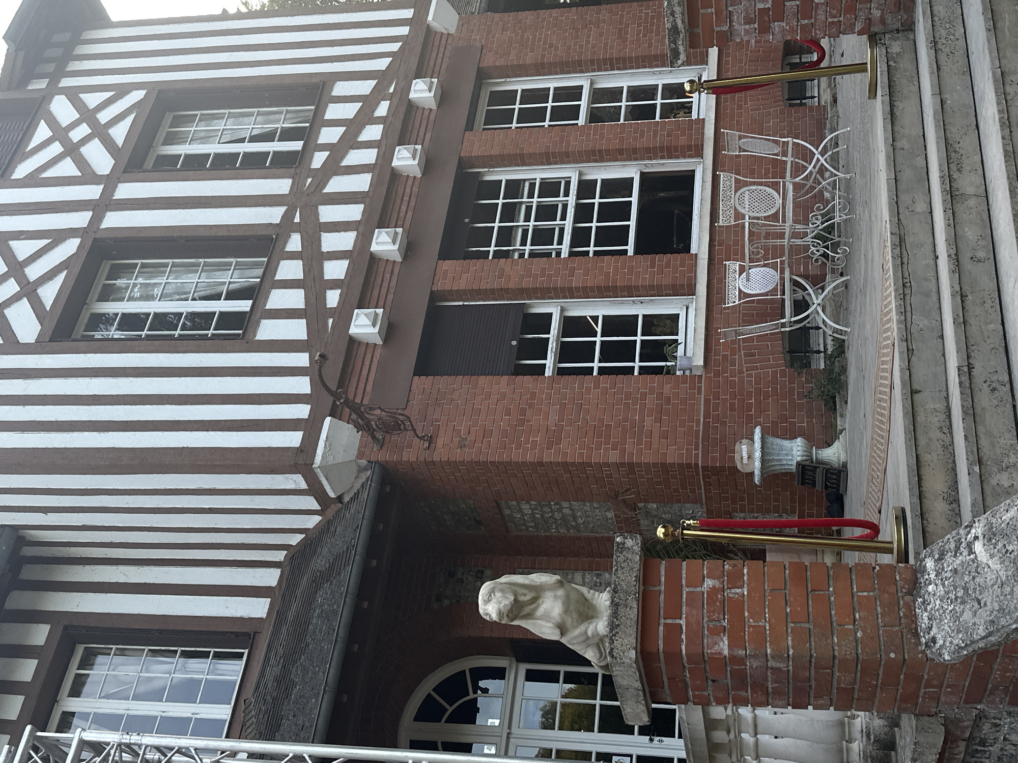 The image shows the exterior of a brick building with a timber-framed upper story, characteristic of Norman architecture, likely located in Saint-Vaast-du-Val, France. The building features multiple tall, multi-paned windows with white frames. A statue of a dog, possibly a Basset Hound, sits on a brick pillar to the left of the entrance. In front of the building, a set of stone steps leads up to a patio area where a white metal table and four chairs are arranged. Red velvet ropes on gold stanchions are present, suggesting a controlled entrance or a VIP area. The overall scene appears to be a daytime shot, with natural light illuminating the facade.