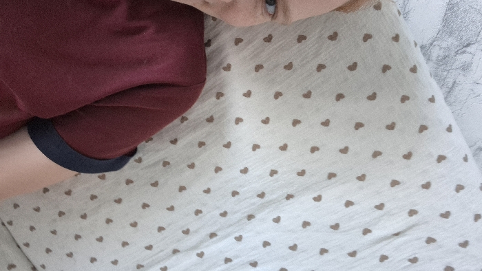 The image displays an indoor scene within the city of Chaikovsky, Russia. A person, identified by a partial view of their face and upper body, occupies the left side of the frame. The visible facial features include a blue-grey left eye, part of the nose, and forehead, with reddish hair visible on the top. The individual is wearing a dark red or maroon short-sleeved shirt with a distinct dark blue cuff on the visible sleeve. To the right of the person, a light-colored fabric, likely a pillow or bedding, is present, patterned with numerous small, evenly distributed light brown heart shapes. A light grey, textured surface, possibly a wall or headboard resembling marble or patterned wallpaper, is visible in the upper left corner behind the person's head.