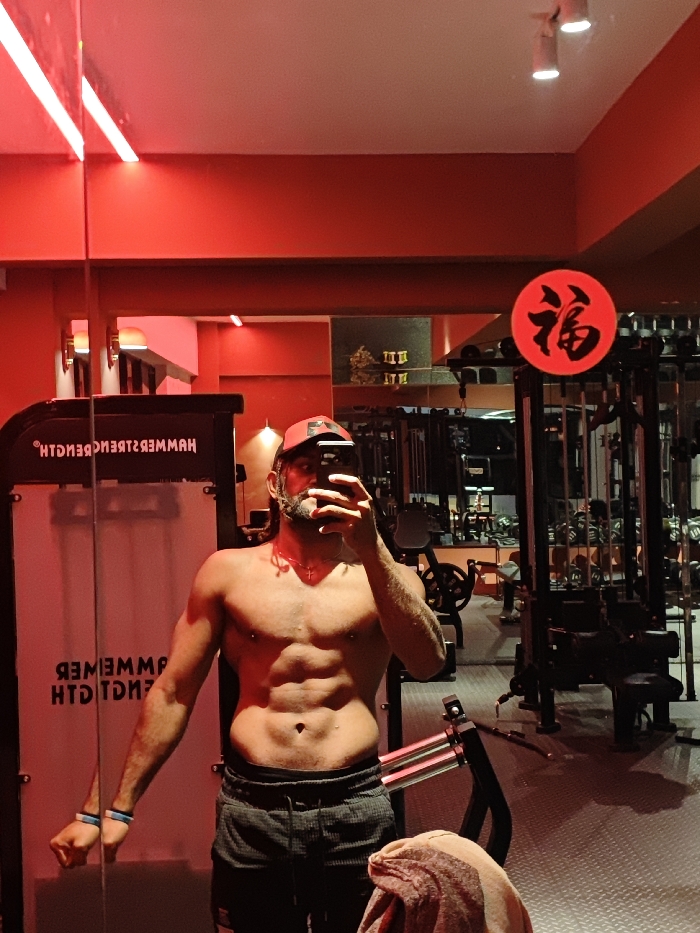 An adult male is depicted taking a mirror selfie inside a gym located in the Chongqing High-tech Zone, China. The scene is dominated by red lighting, illuminating red-painted walls and a gray floor, possibly rubberized. The man, shirtless and displaying a muscular physique with visible abdominal definition, wears dark grey athletic shorts, a red baseball cap, and a red necklace with a small cross pendant. A blue and white wristband is on his left wrist. He holds a black smartphone in his right hand, obstructing part of his face. To the left of the man's reflection, part of a black weight machine is visible with the reversed text "HAMMER STRENGTH BENCH." In the background, other gym equipment and weight racks are visible. A circular red sign featuring the black Chinese character "福" (fú, meaning fortune or good luck) is positioned in the upper right section of the reflection. White spotlights are present on the ceiling.