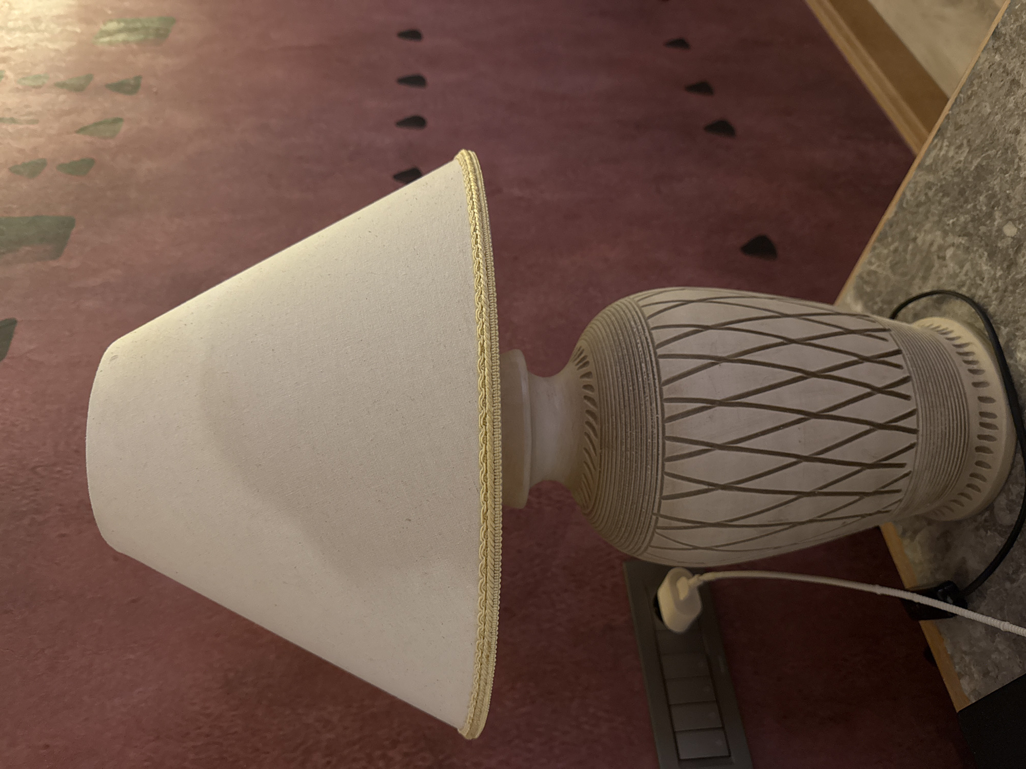 A lamp with a white lampshade and a patterned base sits on a surface in what appears to be an indoor setting. The background features a surface with a repeating triangular pattern. The photograph does not contain sufficient information to determine the location in the city of Kemer, Turkey, or any details about people or actions.
