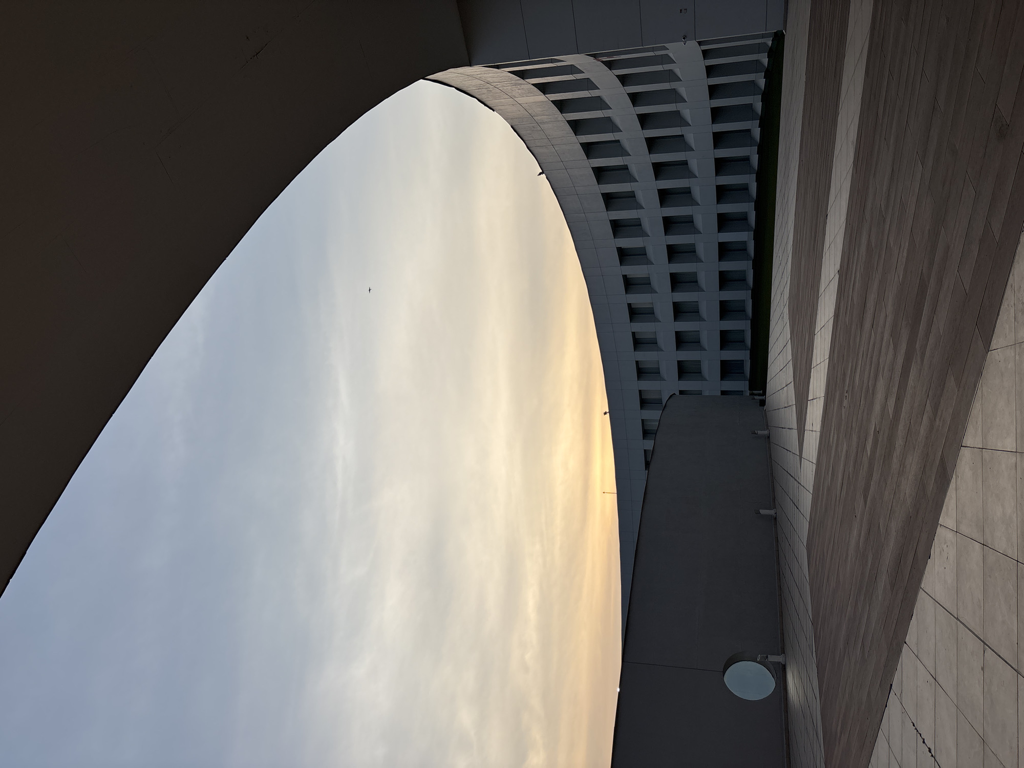 This is an upward-looking perspective of an architectural structure, likely in Mexico City. The sky is visible in the center, with a muted gradient of light blue and yellow hues, suggesting either sunrise or sunset. A building with a repetitive, square-grid facade is on the right, while a textured wall with angular patterns is on the bottom right. The photo seems to be taken from within or under an arch, which frames the sky. A small plane flies across the sky.