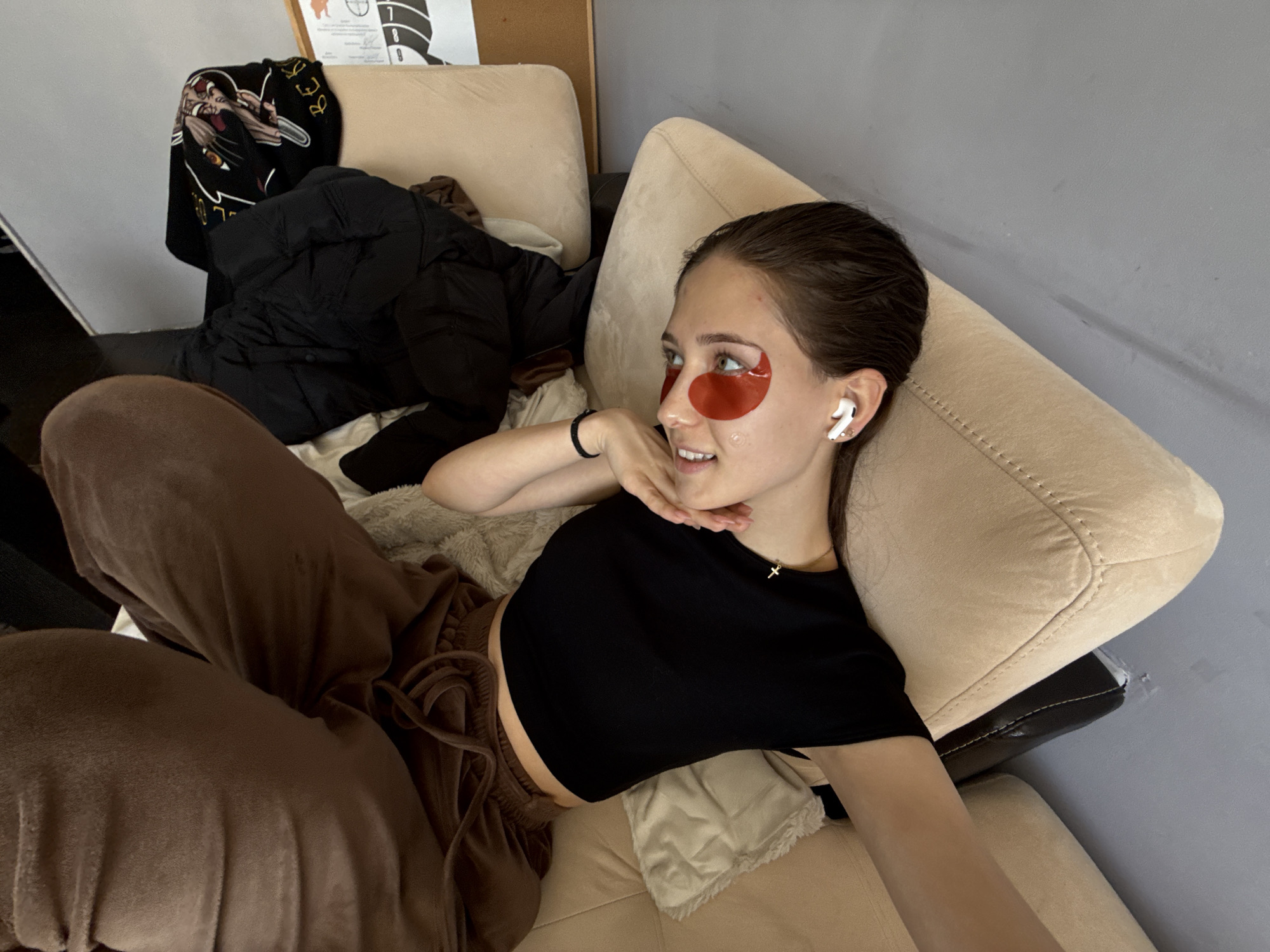 A woman is reclining on a beige couch. She is wearing brown sweatpants, a black crop top, and has red eye masks on. She has earphones in her ears. A puffy black jacket and a black jacket with white print are draped over the couch behind her. A target is on the wall behind the couch. The location is in Sofia, Bulgaria.