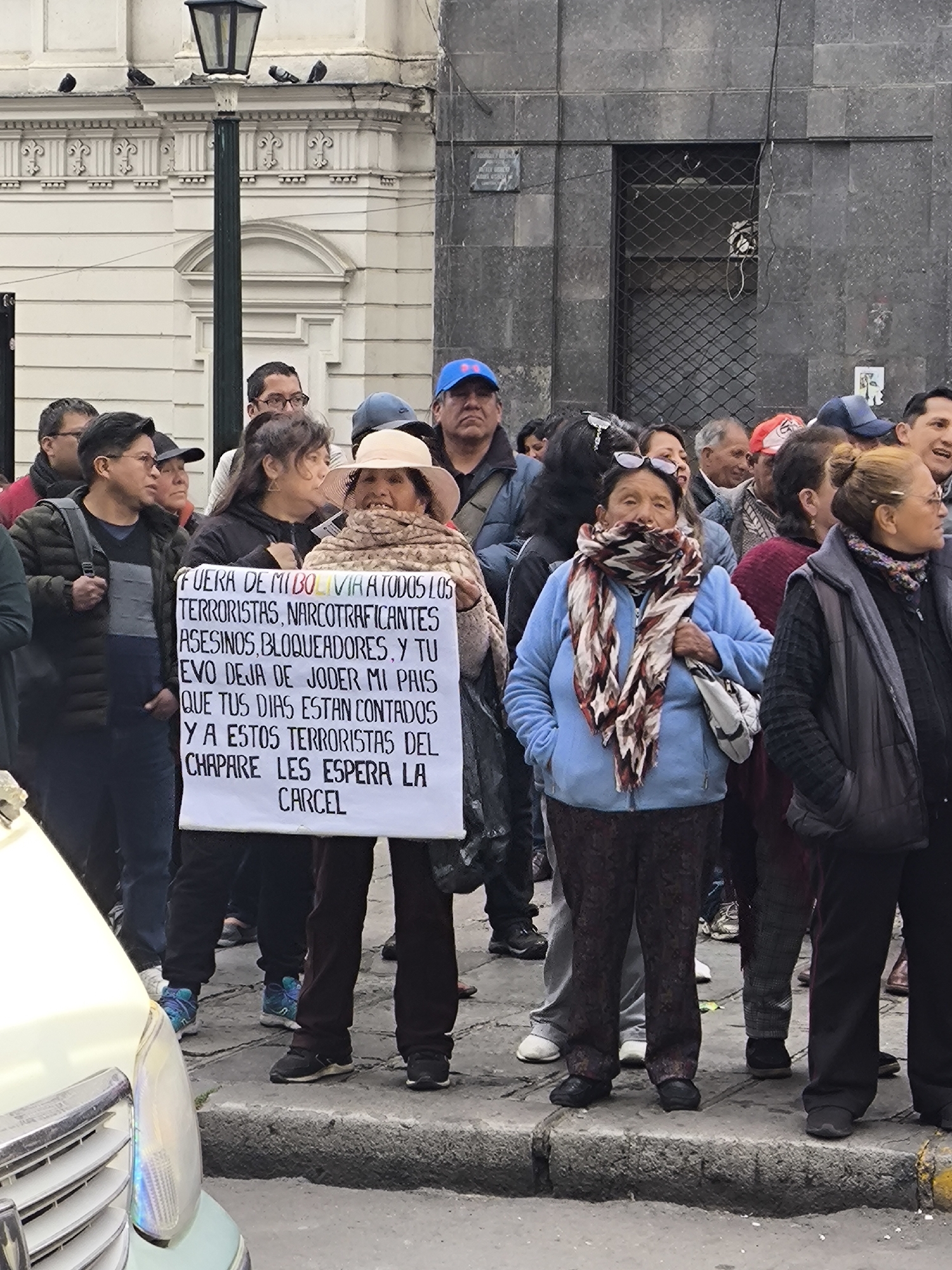 The image depicts a street scene in La Paz, Bolivia, where a group of people are gathered, some holding a large white banner. The banner, held by a woman wearing a light-colored hat and a patterned scarf, contains text written in black ink. The text reads: "FUERA DE MI BOLIVIA A TODOS LOS TERRORISTAS, NARCOTRAFICANTES ASESINOS, BLOQUEADORES, Y TU EVO DEJA DE JODER MI PAIS QUE TUS DIAS ESTAN CONTADOS Y A ESTOS TERRORISTAS DEL CHAPARE LES ESPERA LA CARCEL". This translates to: "OUT OF MY BOLIVIA ALL THE TERRORISTS, NARCOTICS TRAFFICKERS, MURDERERS, BLOCKERS, AND YOU EVO STOP FUCKING MY COUNTRY YOUR DAYS ARE COUNTED AND THESE TERRORISTS FROM CHAPARE ARE WAITING FOR JAIL."
The crowd consists of individuals of various ages and ethnicities, dressed in casual clothing. Some people are looking at the camera, while others are looking in different directions or at the banner. In the background, buildings with architectural details are visible, including a lamppost and a stone-tiled wall. The setting appears to be an urban environment, possibly a city square or a street with pedestrian traffic. The overall mood of the image suggests a protest or demonstration, with the banner conveying a strong political message.