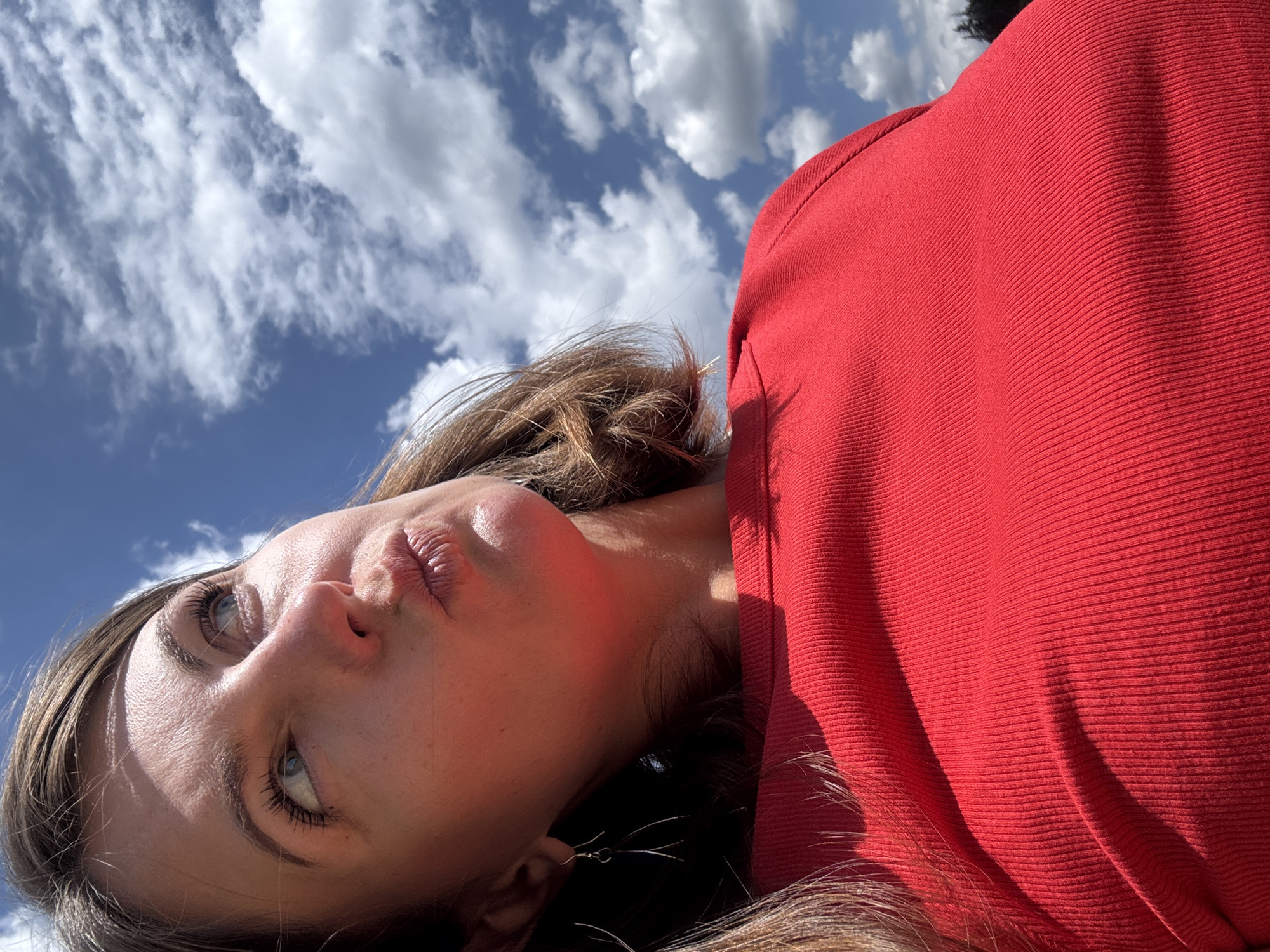The image is a close-up, low-angle selfie of a woman wearing a red ribbed shirt. Her head is tilted back, and she is looking up towards a bright blue sky filled with white, fluffy clouds. Her lips are pursed, and her eyes are open. Her hair is brown and falls around her shoulders. She is wearing a silver earring in her visible ear. The background is dominated by the sky and clouds, suggesting an outdoor setting. There is no indication of the specific location within Coteaux du Lizon, France.