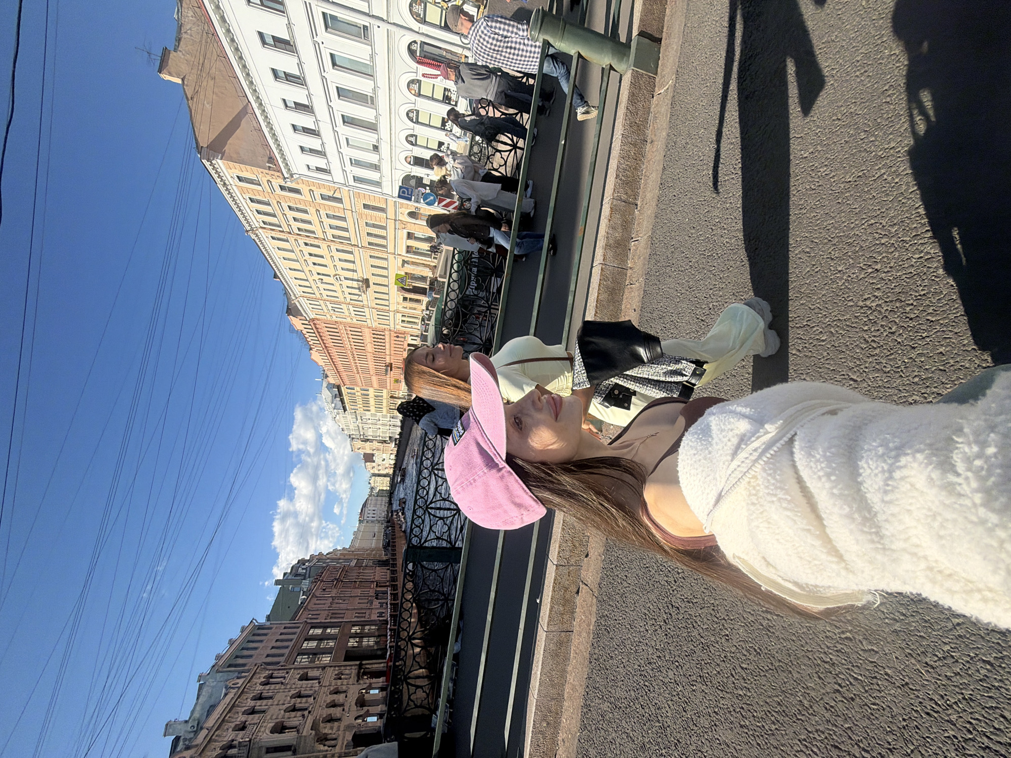 The image depicts a sunny day scene on a bridge in Saint Petersburg, Russia, with a canal visible below and historic buildings lining the waterway. In the foreground, two young women are walking. The woman on the left, facing to the right and slightly away from the camera, wears a pink baseball cap, a brown tank top, and a white fuzzy jacket. She has long brown hair and is looking to her right. The woman next to her, slightly behind and to her right, faces forward with her eyes closed. She wears a pale green top and loose-fitting pants, with a black and white patterned jacket draped over her arm.
In the background, other pedestrians are visible crossing the bridge. The canal below is dotted with boats, suggesting it is a popular tourist area. The architecture of the buildings along the canal is typical of Saint Petersburg, with multi-story facades in various colors, including orange, yellow, and white, featuring numerous windows and decorative elements. The sky is clear blue with a few white clouds, and the sun casts long shadows on the asphalt bridge surface. The scene captures a vibrant urban atmosphere during daylight hours.
