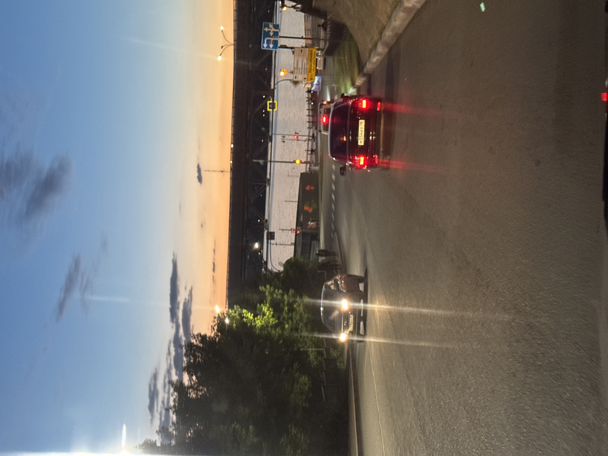 The image captures a twilight scene on a city street in Izhevsk, Russia, likely near the Izh River. The sky exhibits a gradient of colors, from a deep blue at the top to a warm orange and yellow near the horizon, indicating sunset or sunrise. Several vehicles are visible on the asphalt road, with their headlights and taillights illuminated.
In the foreground, on the right side of the road, a black sedan is traveling away from the viewer. Its license plate reads "M554KY 18". The car's red taillights are prominent, casting reflections on the wet road surface. Further down the road, other vehicles are also present, their lights a blur in the low light conditions.
To the left of the road, a dense cluster of trees stands tall, their dark foliage contrasting with the illuminated sky. The trees are partially lit by streetlights or other artificial light sources, with some leaves appearing yellowish.
In the background, a wide river is visible, reflecting the sky's colors. A large bridge spans the river, with structural elements and lights visible against the twilight sky. Traffic signals and streetlights are scattered throughout the scene, adding to the urban ambiance. A pedestrian crossing signal is also visible on the right side of the road, suggesting an intersection or pedestrian crossing area. The overall impression is of a busy urban environment at dusk.