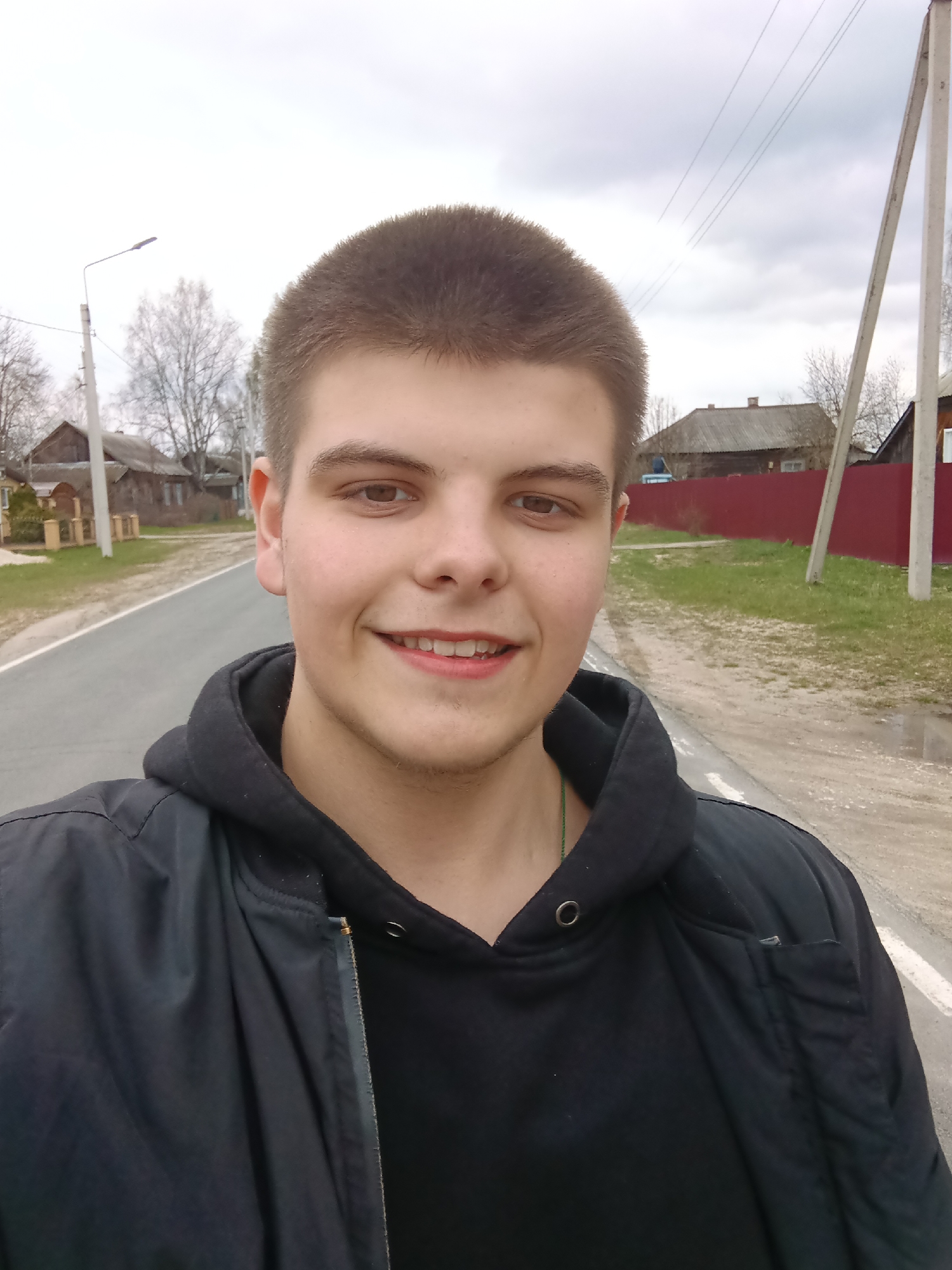 A young man with short brown hair and a black jacket stands smiling on a paved road. The location is Красный Маяк, Russia. The background shows houses, trees, a street light, and an overcast sky. A red fence is visible in the background, along with utility poles and wires. Patches of grass and puddles are present alongside the road.