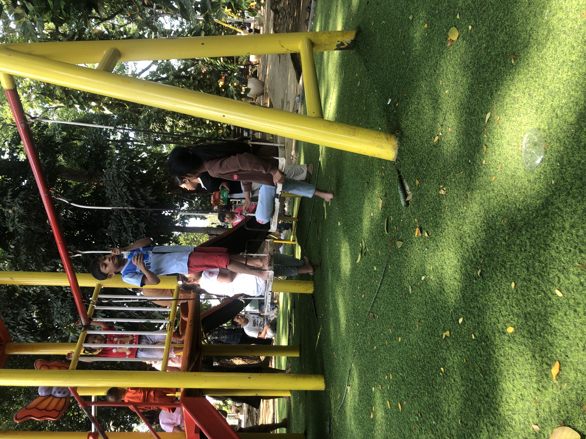 The image depicts a playground in Pasuruan, Indonesia. A yellow and red metal swing set is the central element, with green artificial turf covering the ground, scattered with yellow leaves. Several children and adults are present. In the foreground, a young boy in a blue shirt and red pants stands near a swing, holding a small plastic bottle. To his right, a girl in a brown shirt and blue jeans sits on a swing, her bare feet dangling. Behind them, other individuals, including adults and children, are visible in the background, some standing and others sitting. The playground structure features yellow poles and a red railing. Lush green trees and foliage surround the playground, suggesting an outdoor setting.