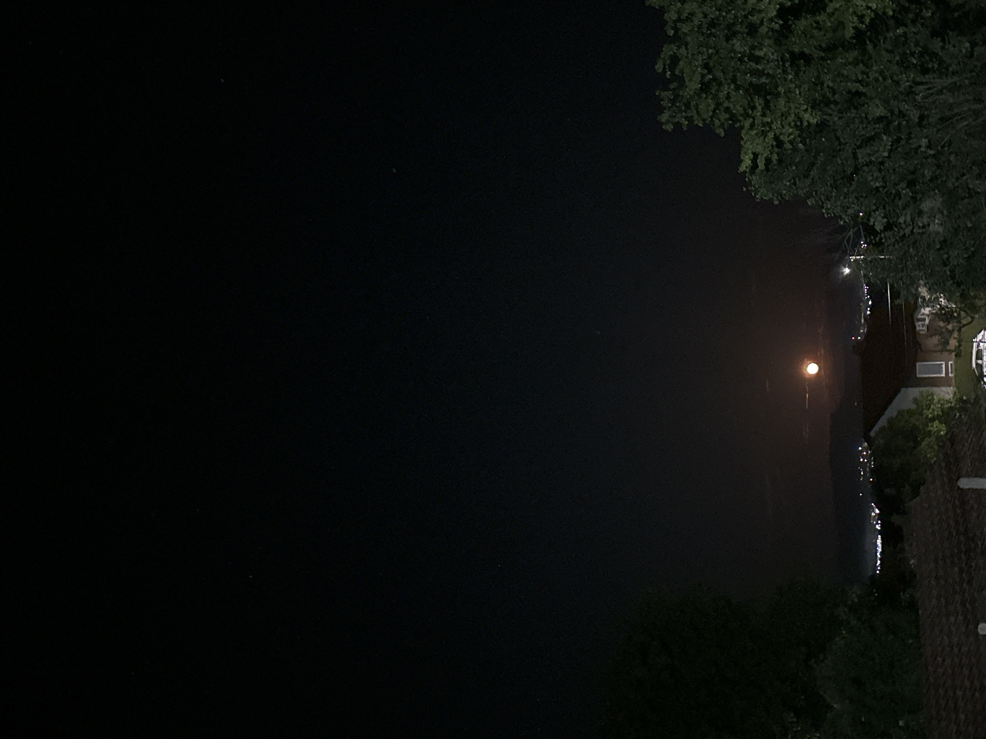 The image captures a tranquil night scene, likely taken from an elevated position overlooking a body of water in the Kormiansko area of Bulgaria. The predominant feature is a dark, clear night sky, which fills most of the left and upper portions of the frame.
A large, bright, and distinctly round, yellowish-orange moon is prominently visible in the upper mid-right of the image, illuminating parts of the scene. Below the moon, a dark expanse of water stretches out, reflecting numerous small, glittering artificial lights from what appears to be a distant shore or settlement. These reflections create sparkling streaks across the water's surface, indicating calm conditions. The faint outlines of a landmass or hills can be discerned in the far distance, beyond the line of lights.
In the foreground, along the right edge of the frame, dark green foliage from trees partially obscures the view. Below these trees, sections of a dark, tiled roof and parts of what appear to be building walls are visible, suggesting a residential structure close to the vantage point of the photograph.
The scene conveys a peaceful and serene atmosphere, with no people or specific activities depicted. The time of day is clearly night, under a cloudless sky. No visible text is present in the media file.