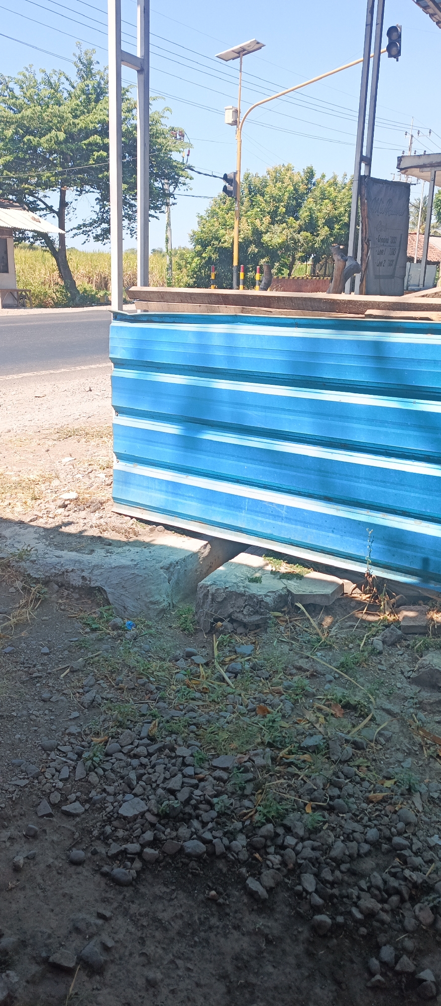 The image depicts an outdoor scene on a sunny day, likely in Jatisari, Indonesia, near a road. In the foreground, there are scattered rocks and dry vegetation on the ground, with a section of blue corrugated metal siding leaning against a concrete structure. Behind the siding, a paved road is visible, with a field of tall, dry grass or stalks to the left. Further back, a light pole with a solar panel and traffic lights stands against a clear blue sky. Trees with dense green foliage are present in the middle ground, and a simple building with a white roof is partially visible on the left. A gray sign with some text, including what appears to be a price list, is mounted on a structure on the right side of the image. The overall impression is of a roadside construction or storage area in a rural or semi-rural setting.