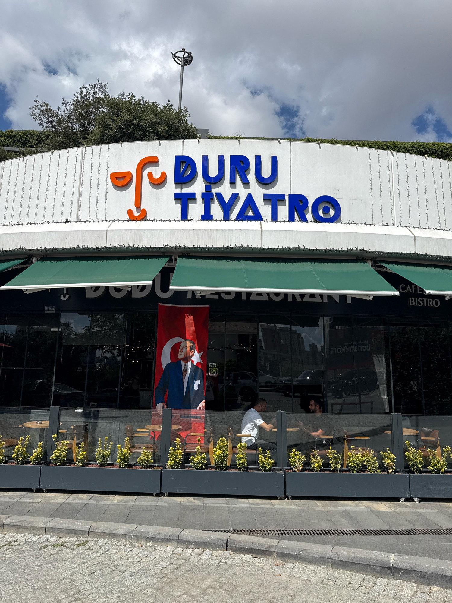 An outdoor medium shot captures the facade of a building with "DURU TİYATRO" signage in blue letters. An orange abstract logo is positioned to the left of the text. The building features a white ribbed exterior wall adorned with string lights. Below the signage, green awnings extend over large glass windows. A large banner displaying a portrait of a man in a suit against a red and white background is hung in front of the windows. Inside, visible through the glass, are tables and chairs, and two men are seated at separate tables. A planter box filled with small green plants runs along the base of the building's exterior. The scene is set on a paved street with a curb in the foreground. The sky above is partly cloudy.