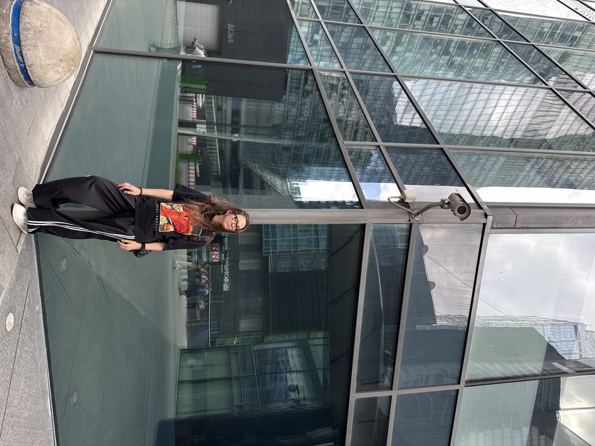 A woman wearing glasses and a graphic t-shirt stands on a grey tiled surface next to a large glass wall. She is wearing black pants with white stripes and white sneakers. To the left of her is a round concrete object with a blue stripe. Behind the glass wall, people are reflected, and a sign with Russian text is visible. Above the glass wall, a modern building with many reflective glass windows is visible. The location is Khimki, Russia, based on the text on the sign. A security camera is mounted above the glass wall.